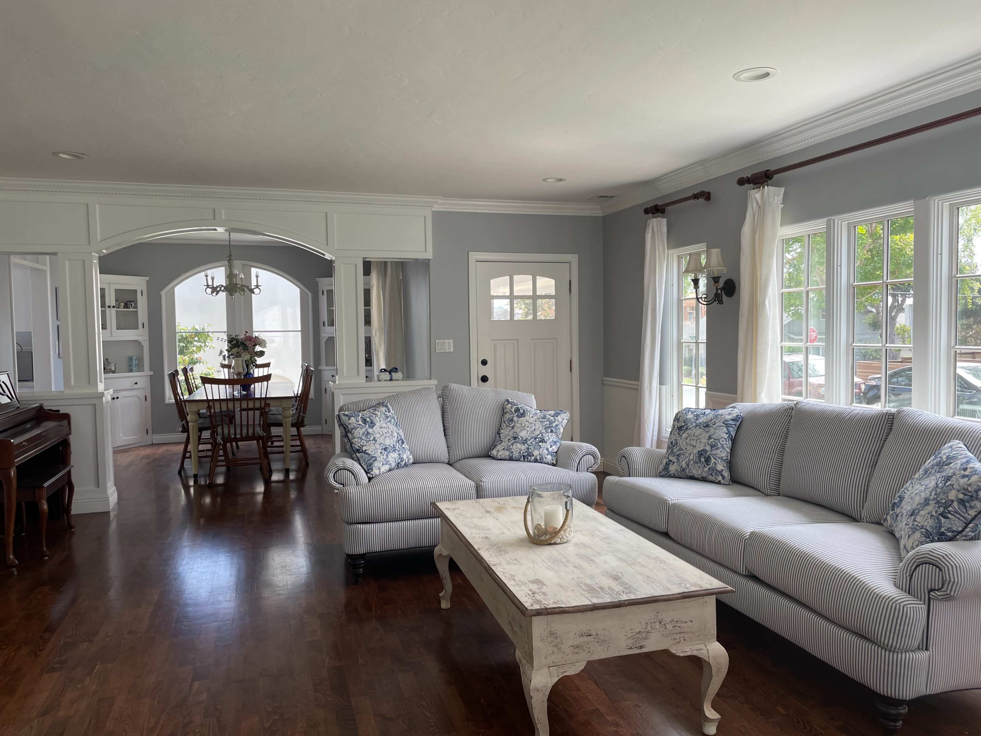 A light-filled living room features a gray striped sofa and armchairs, a wooden coffee table, and a dining area visible through an archway.
