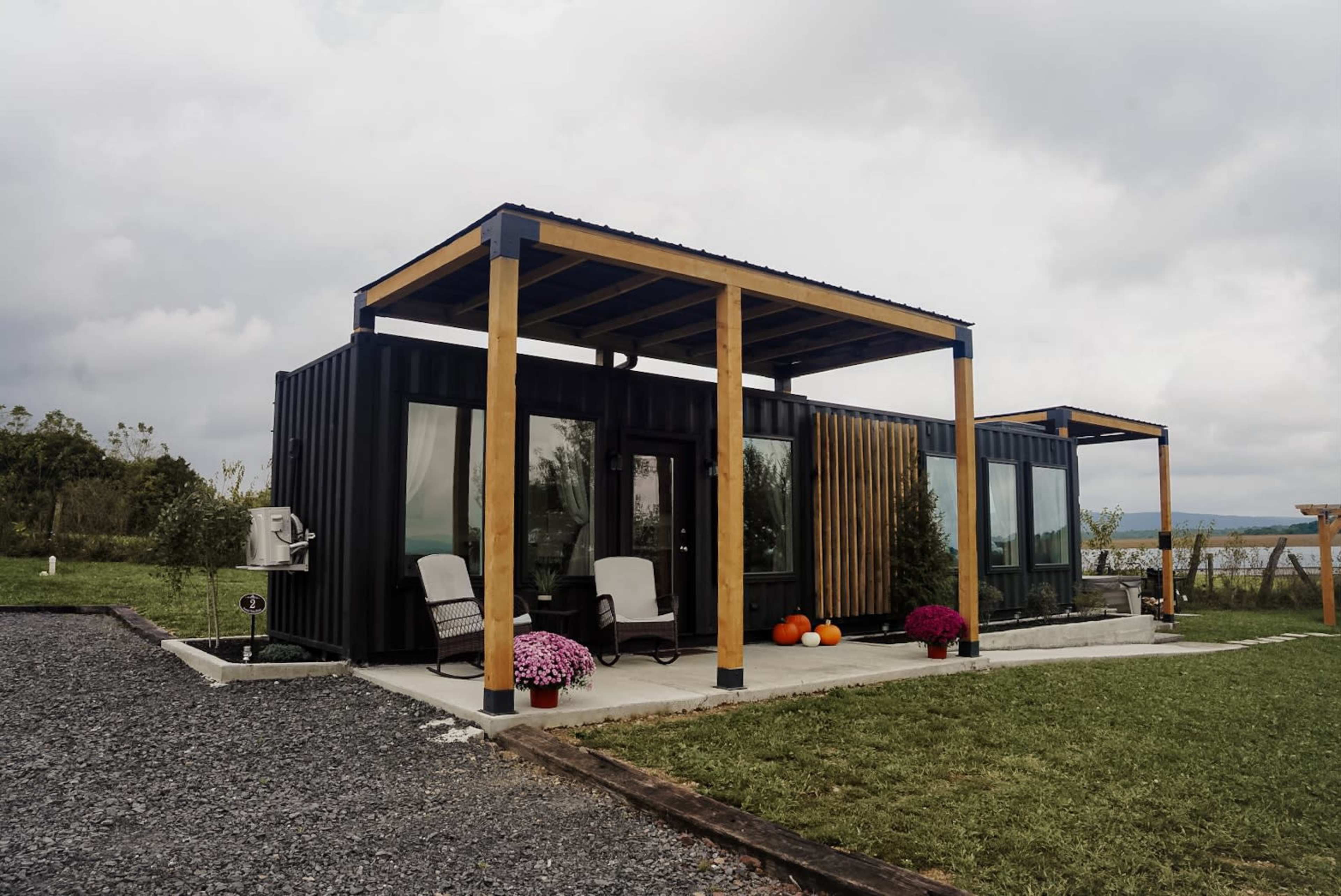 A modern black shipping container house features glass doors, wooden beams, and colorful planters in a landscaped area.