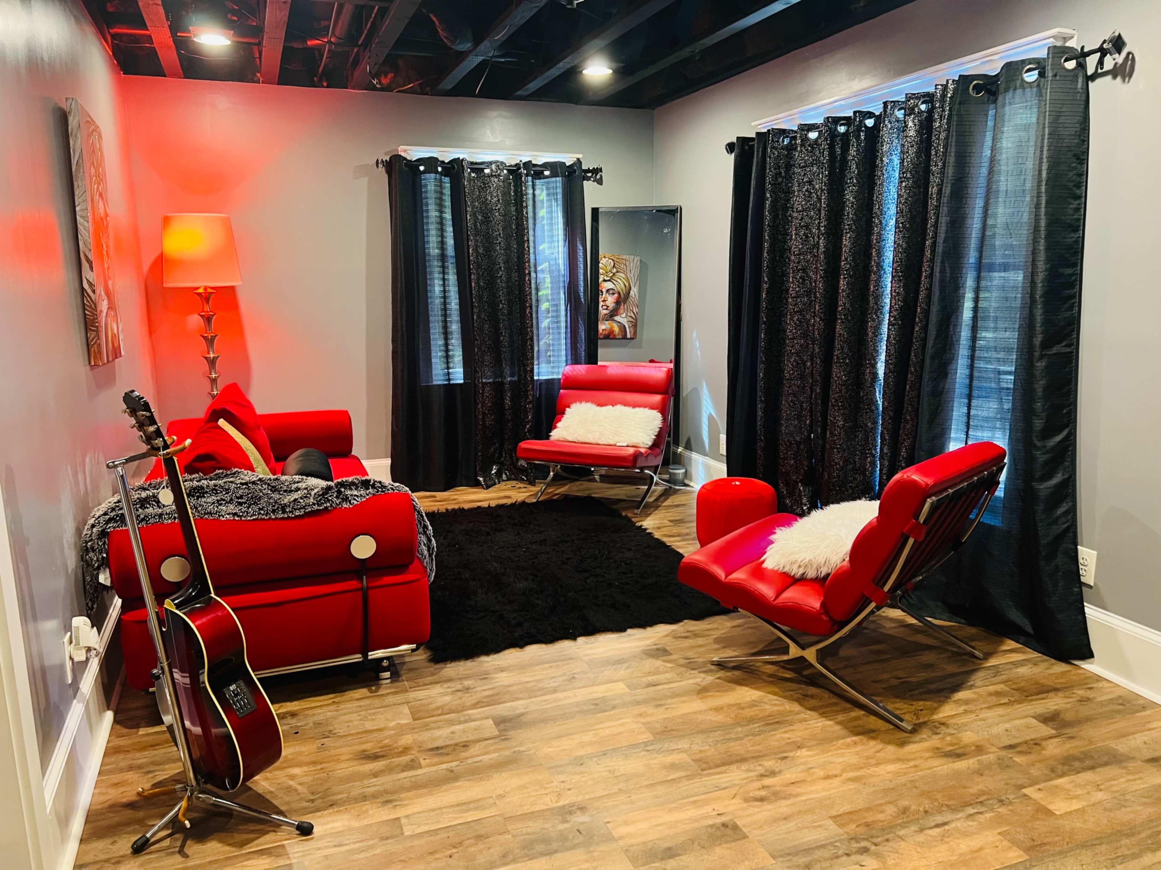 The image shows a modern lounge area with red furniture, a black rug, and dark curtains, featuring a guitar resting against a couch.
