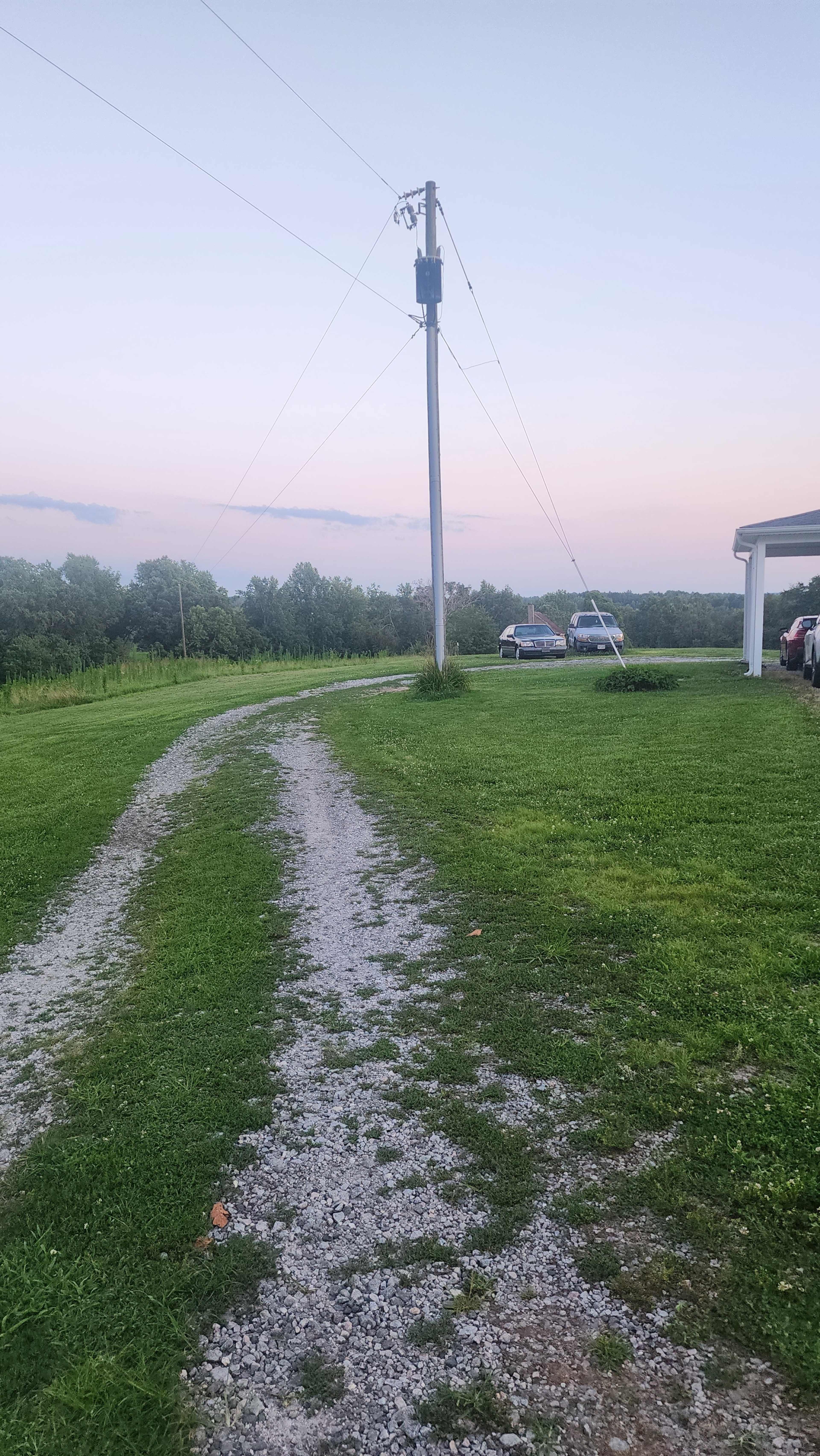 A gravel driveway winds through green grass towards two parked cars near a utility pole, with trees and a sunset sky in the background.