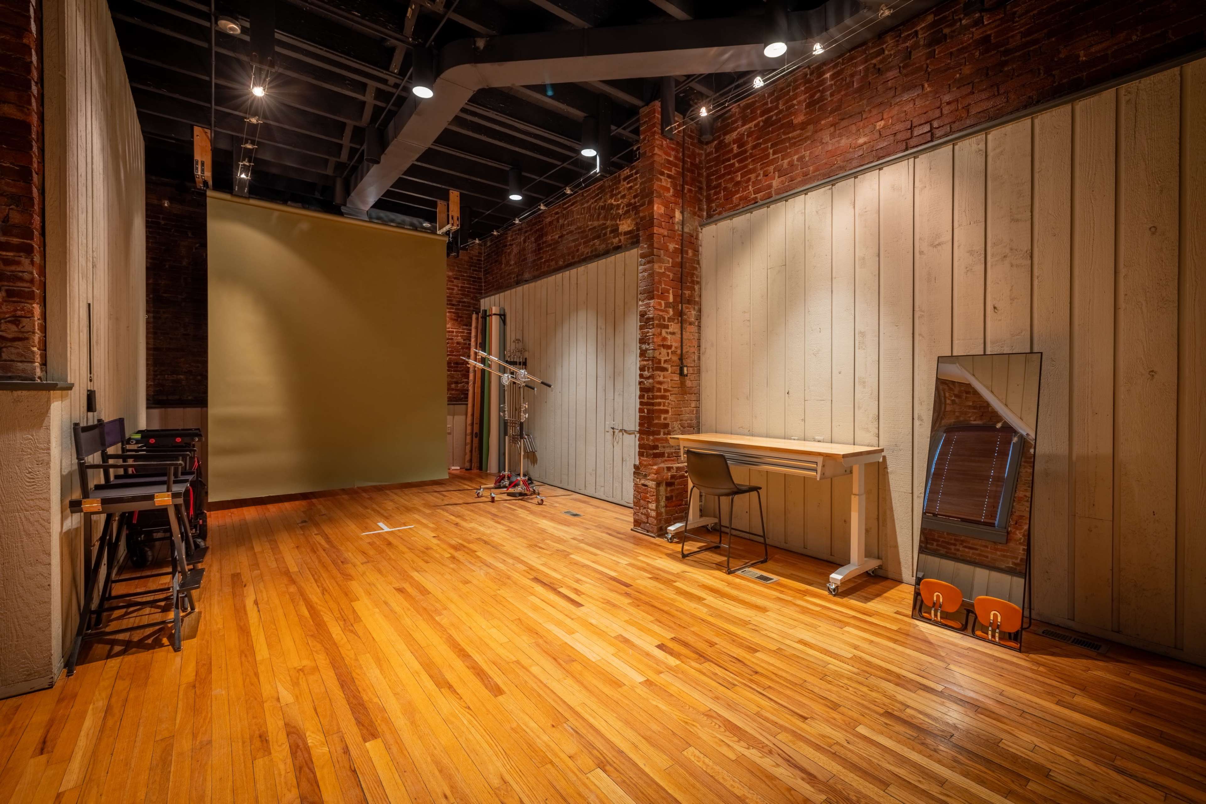 The image shows a spacious, empty studio with exposed brick walls, wooden flooring, a simple desk, a chair, and a large green backdrop against one wall.
