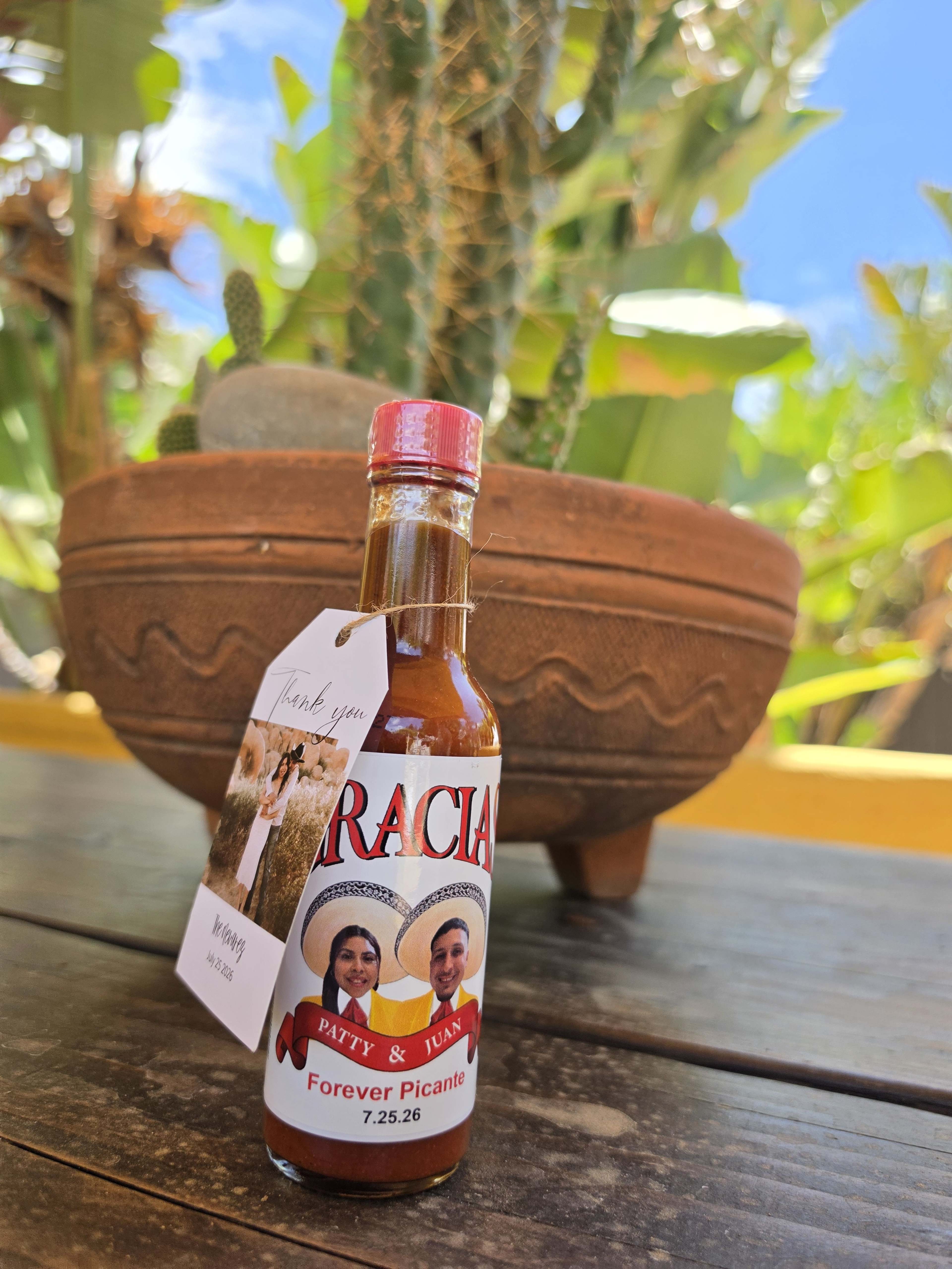 A bottle of hot sauce labeled "Gracia" with a tag reading "Thank you" sits on a wooden table in front of a potted cactus.