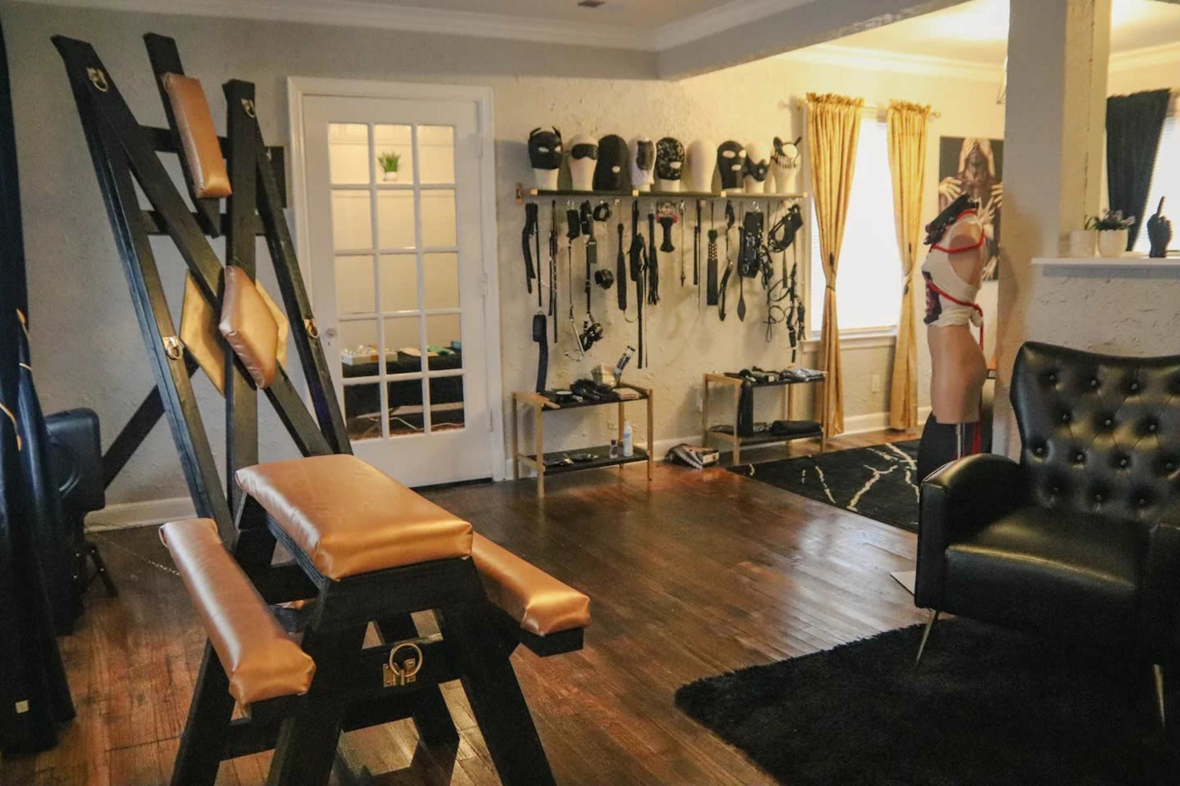 A dimly lit room featuring a black leather chair, a wooden apparatus with padded surfaces, and various BDSM equipment displayed on the walls.
