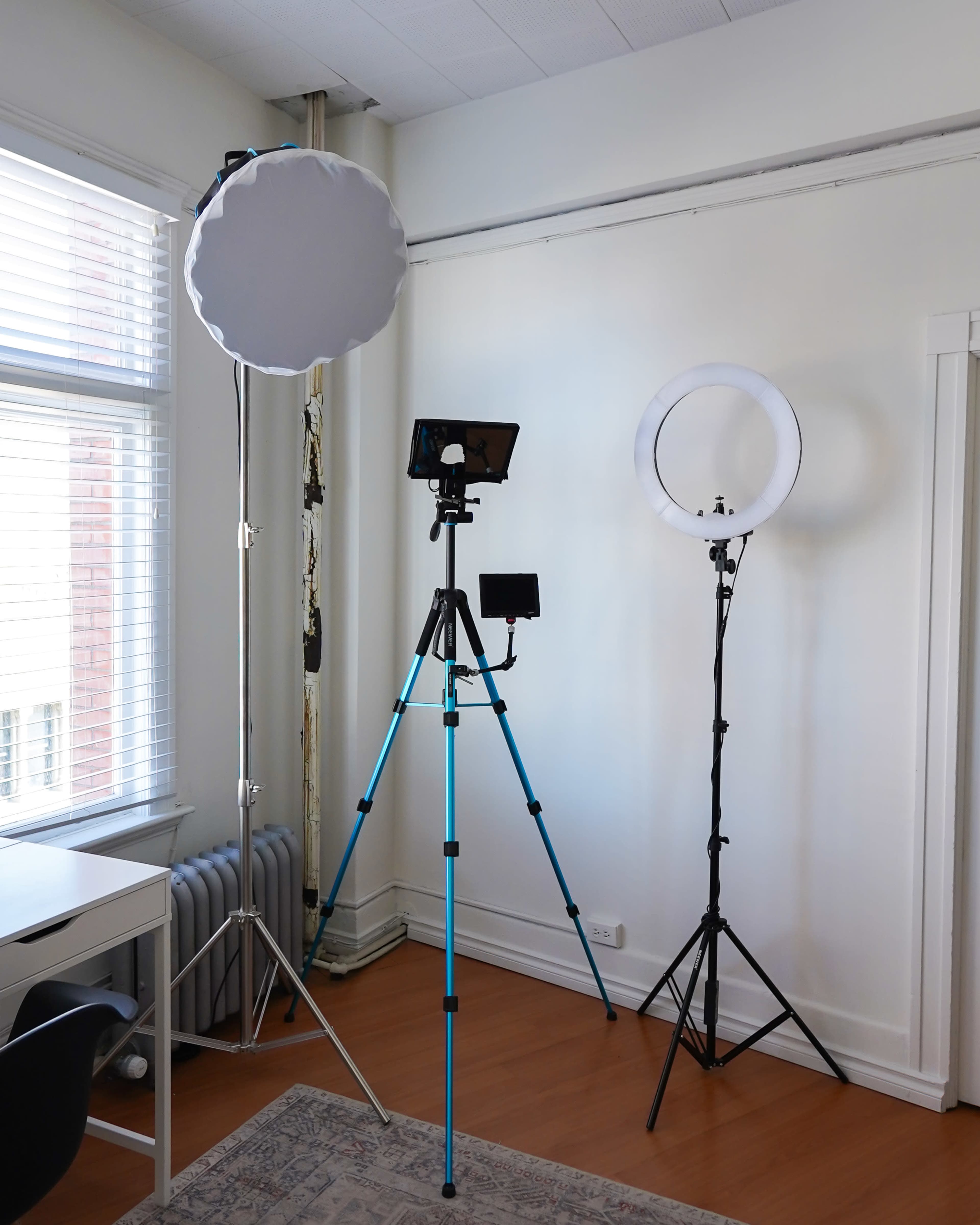 A bright room with large windows, featuring two tripods with lighting equipment and a desk with a chair.