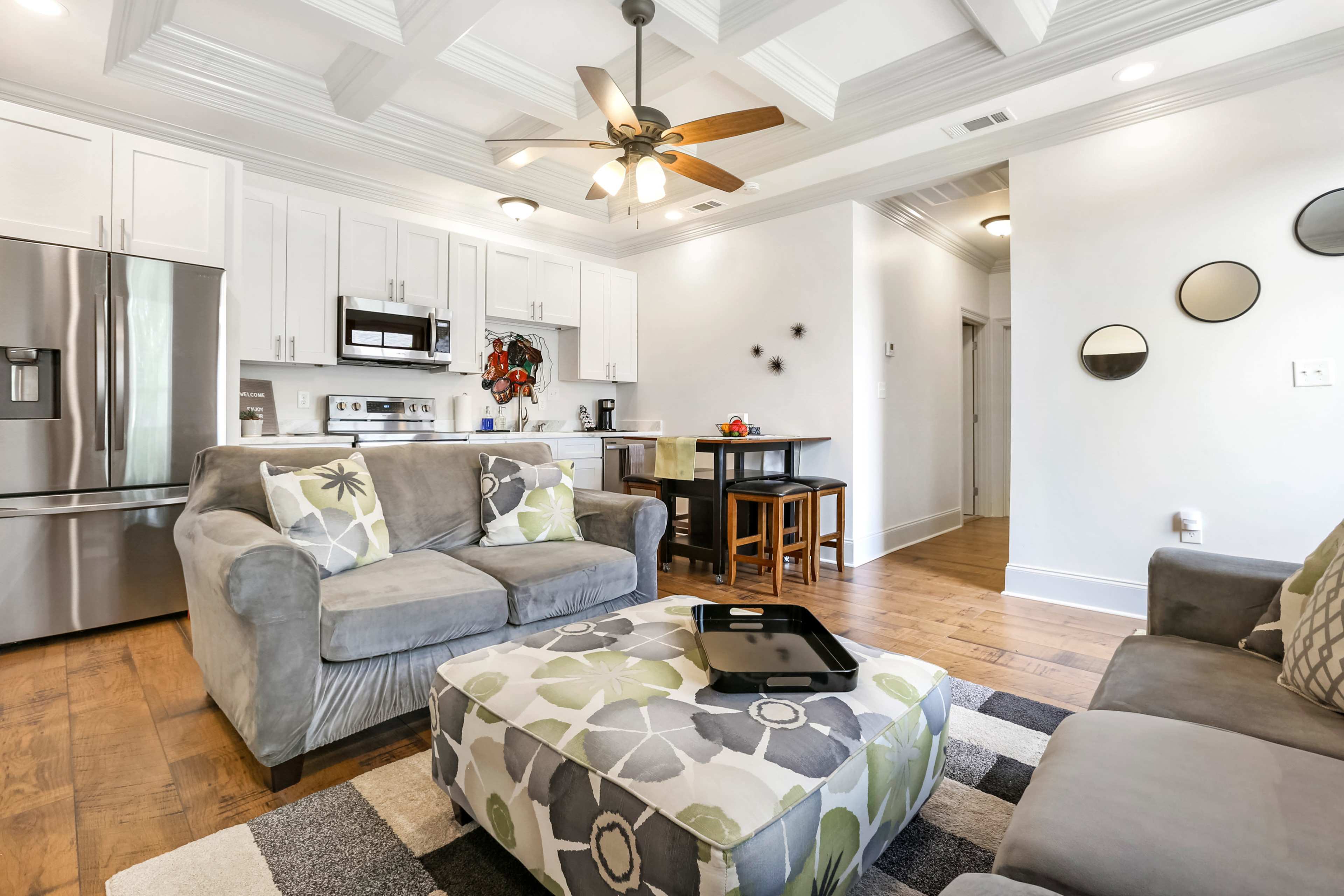 The image shows a modern living room with a gray couch, a patterned ottoman, a kitchen area in the background, and a ceiling fan overhead.
