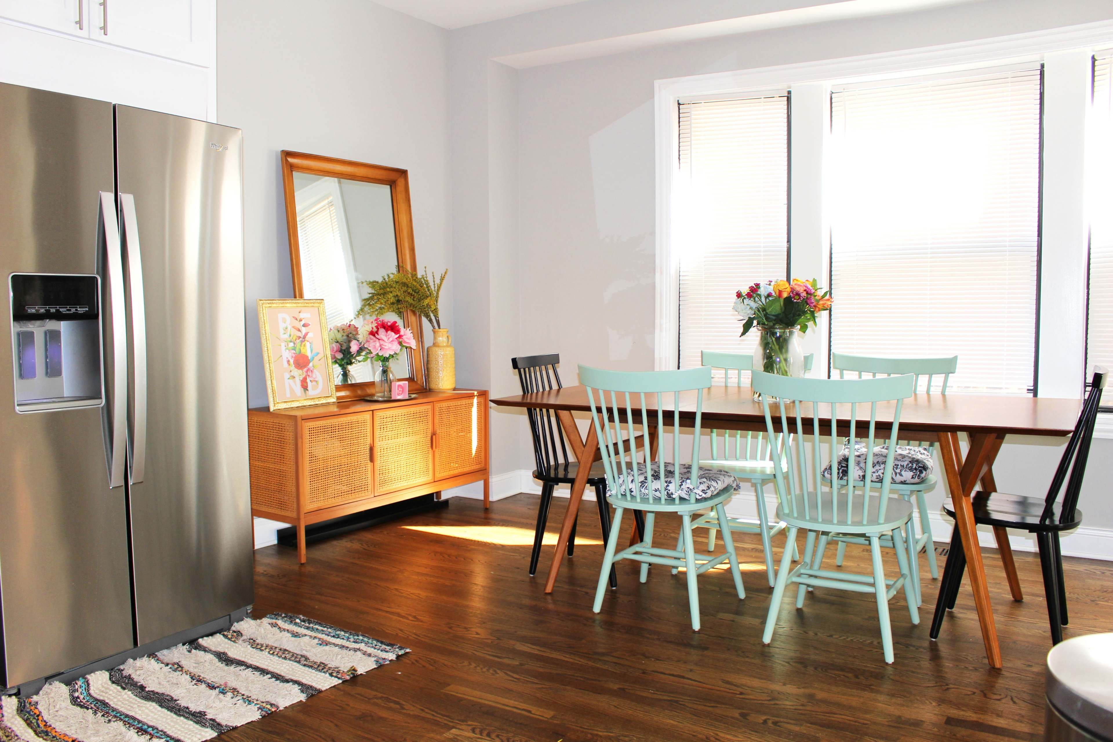 A modern kitchen scene features a stainless steel refrigerator, a wooden dining table surrounded by chairs, and a large mirror on the wall, with natural light coming through the windows.