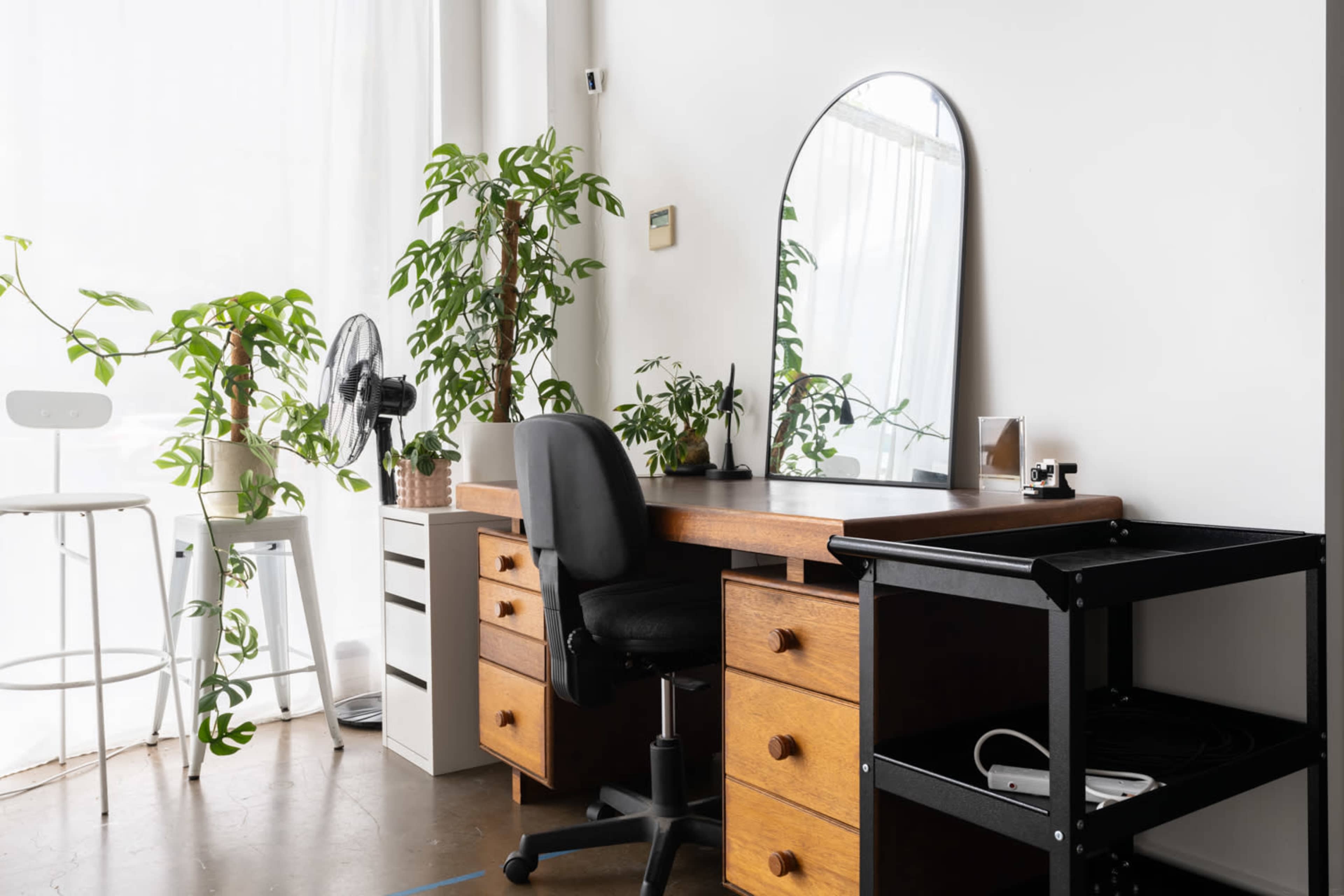 A wooden desk with a large mirror and a black chair surrounded by houseplants in a bright room.