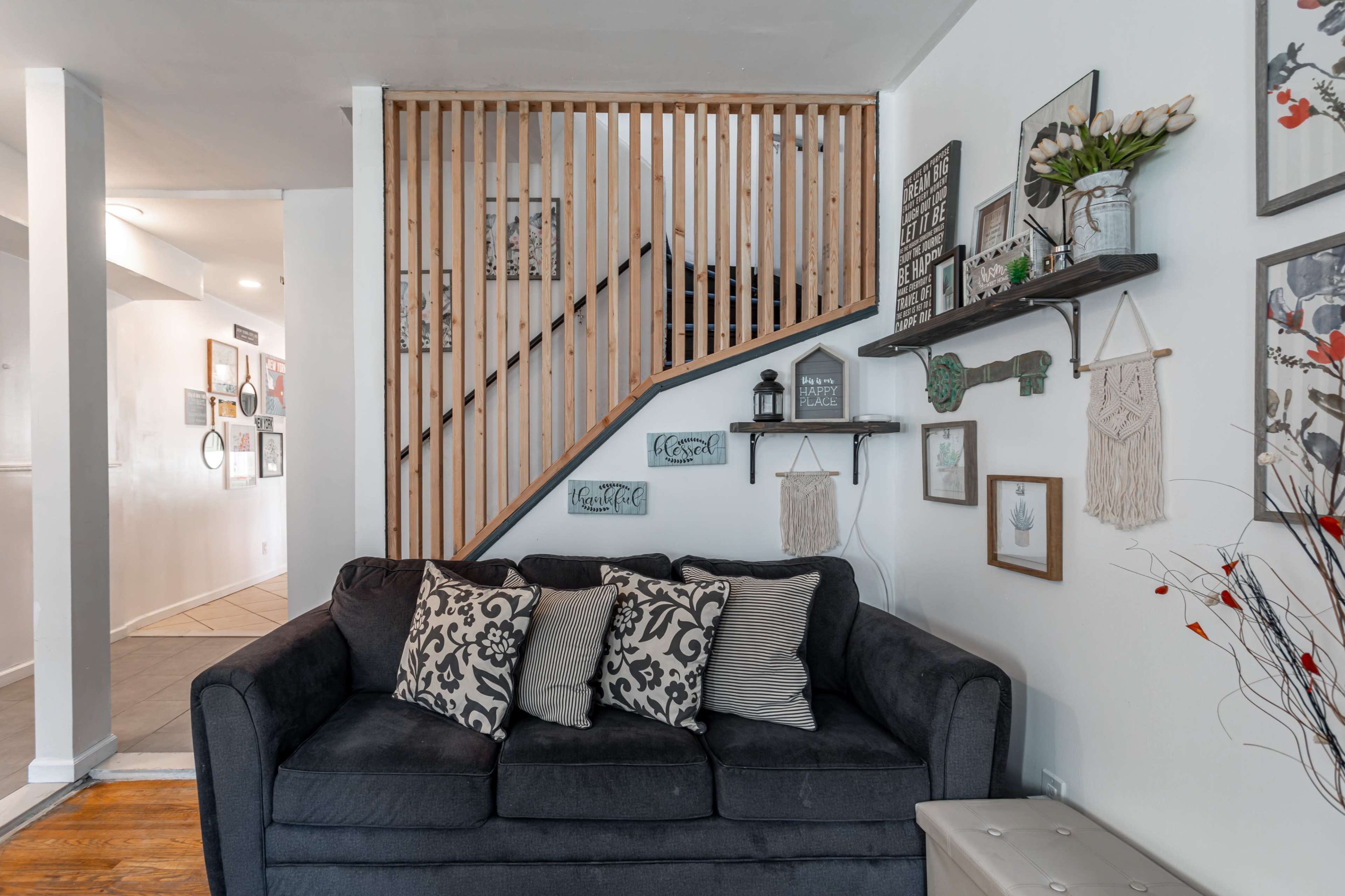 A black couch with decorative pillows is positioned against a wall featuring shelves and framed artworks, next to a wooden staircase.