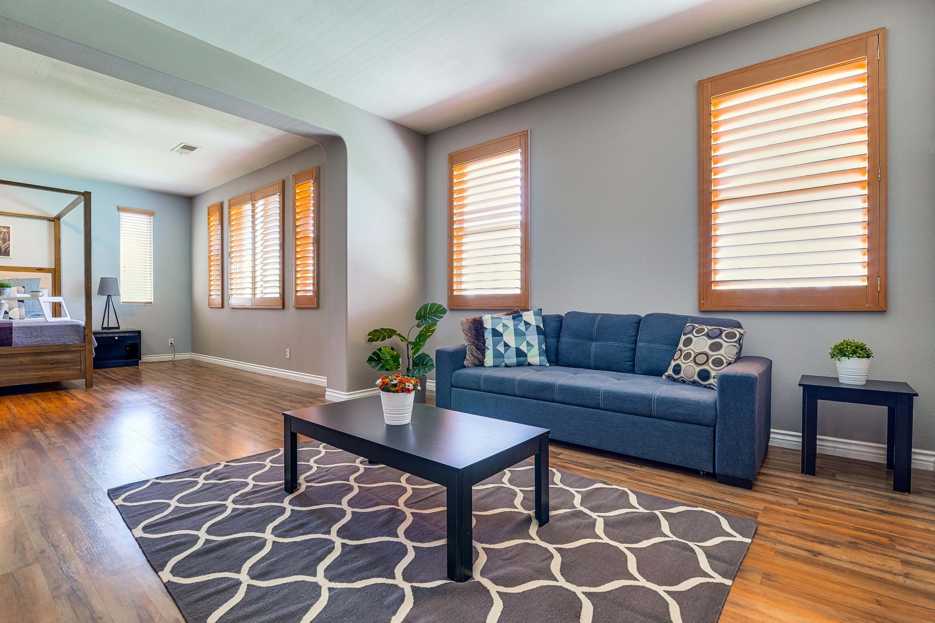 A living room with a blue couch, a coffee table on a patterned rug, and large windows with slatted blinds.