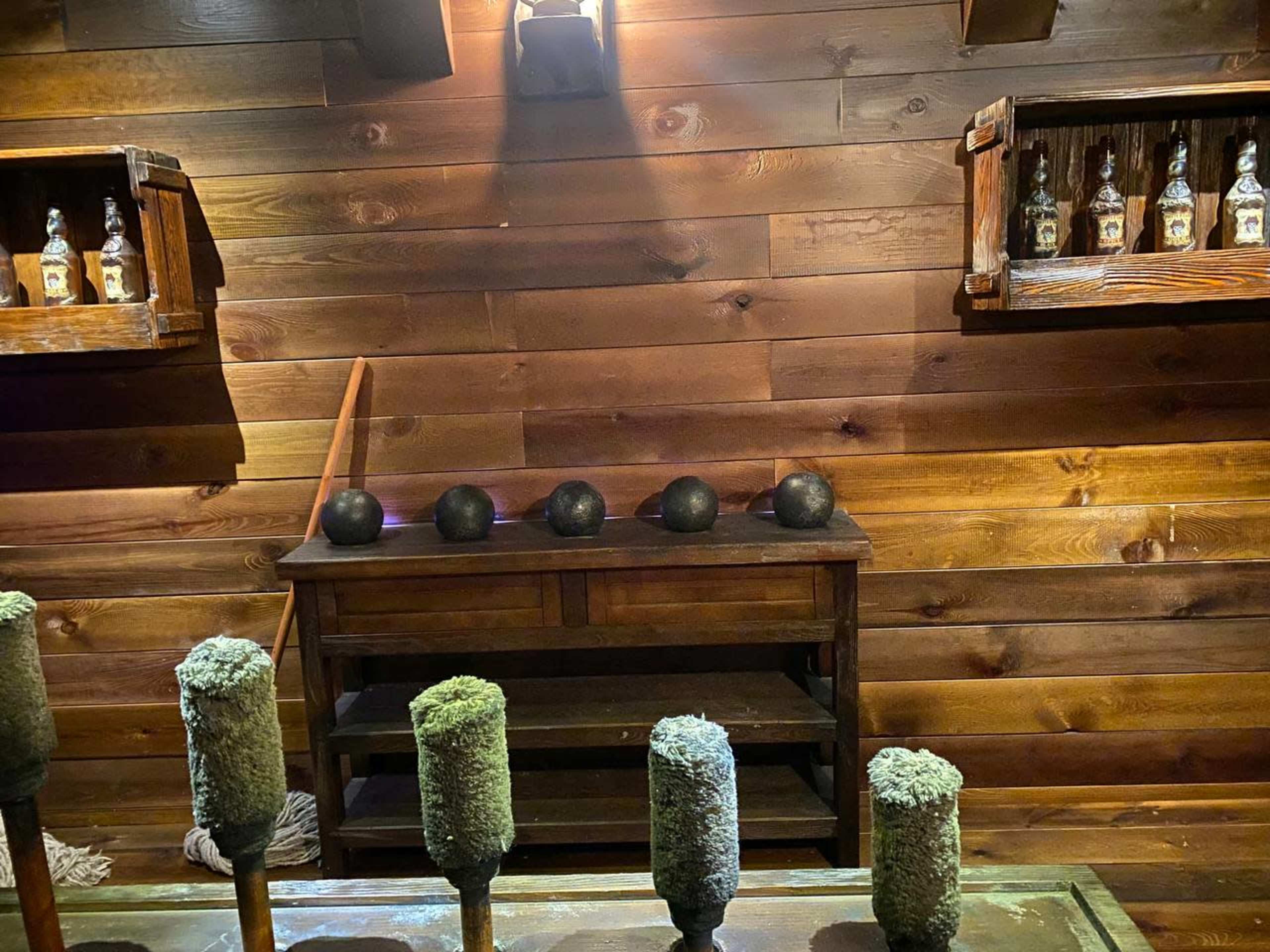 The image shows a wooden interior with shelves containing bottles, a table with a few dark spheres arranged in a row, and bottles on display in the background.