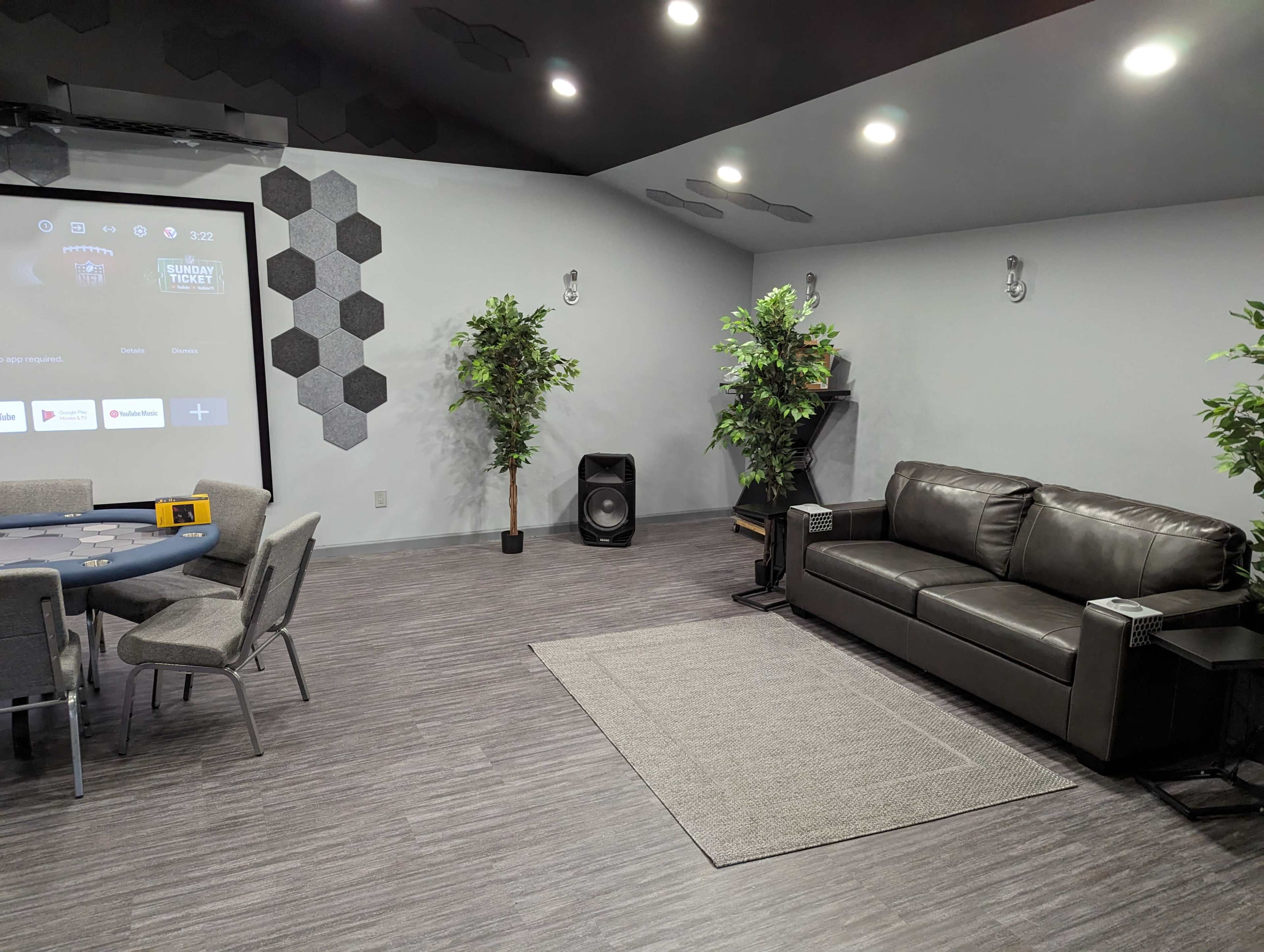A modern room featuring a poker table, a grey sectional sofa, two artificial plants, and a projector screen on the wall.