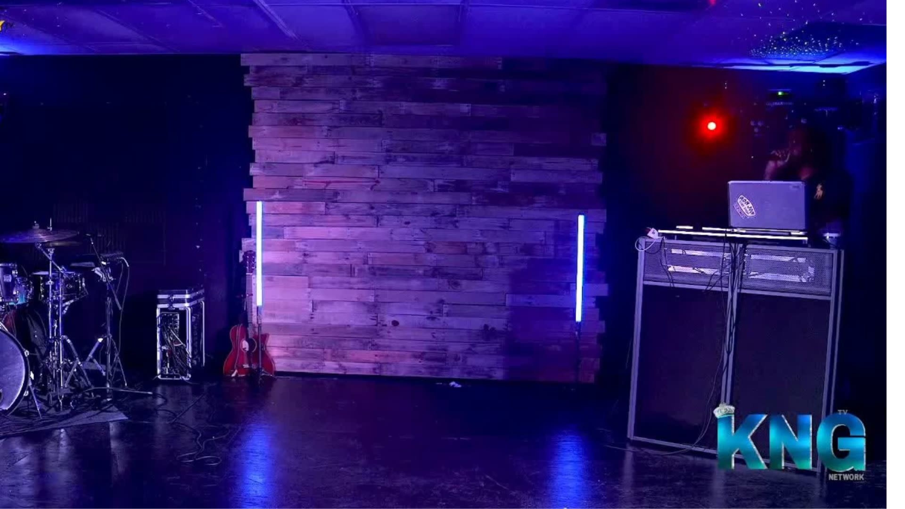 The image shows a performance space featuring a wooden wall, a guitar, a drum set, and sound equipment, illuminated by colored lights.