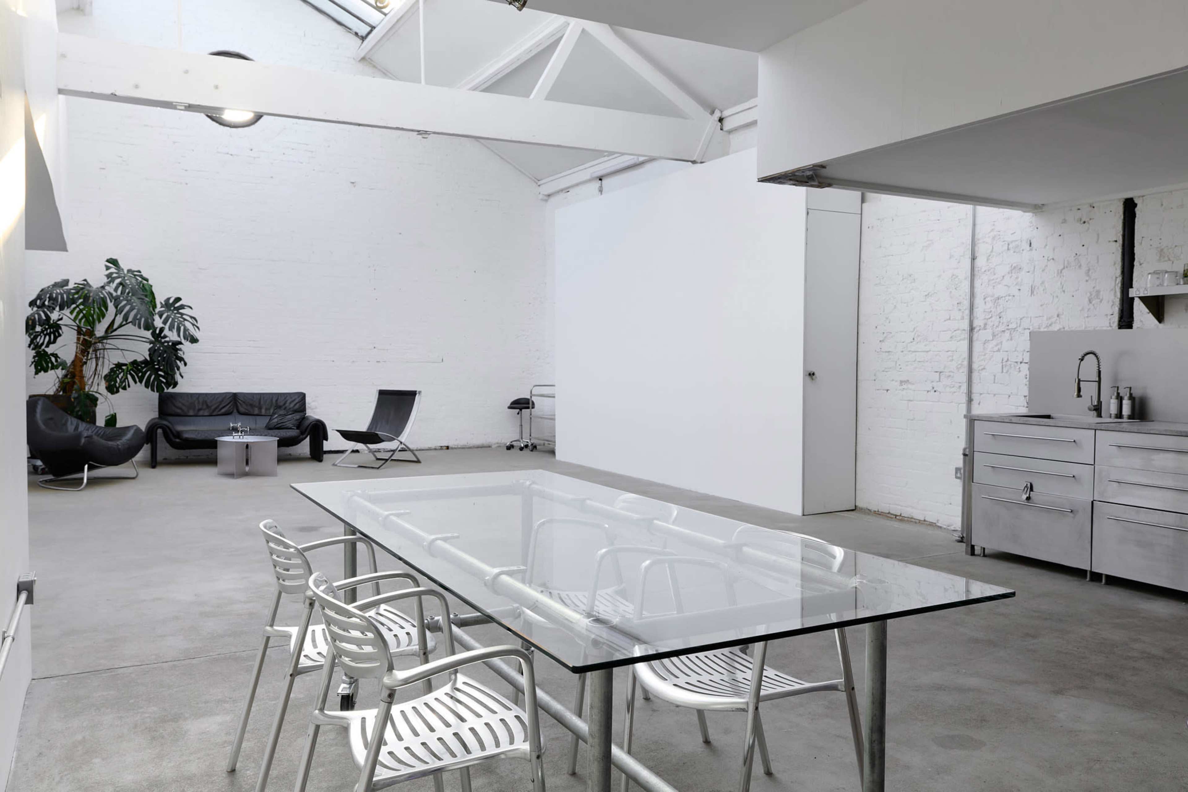 The image shows a minimalist interior space featuring a glass dining table with metal chairs, a sofa, and a potted plant against a white brick wall.