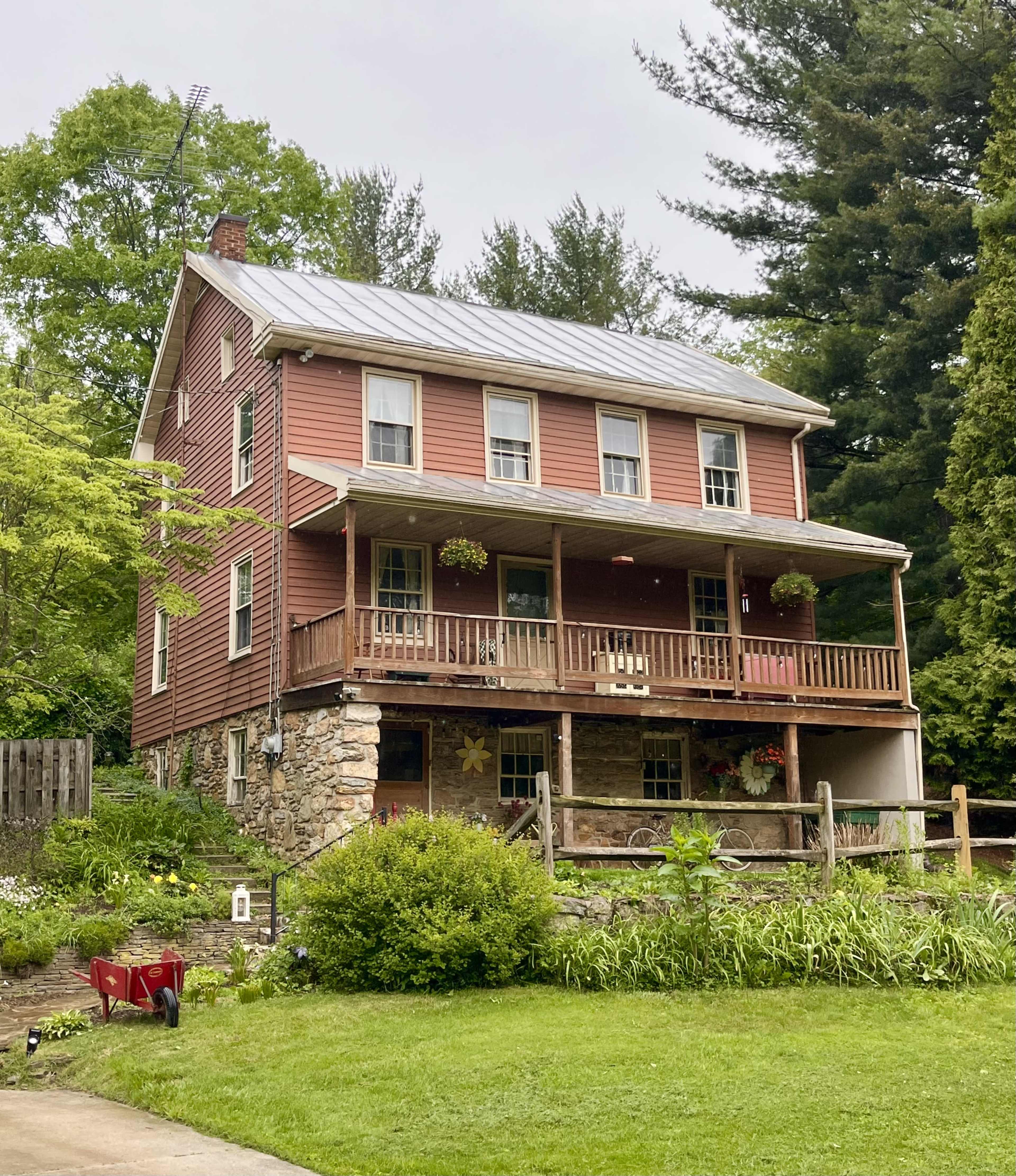 A three-story house with a red wooden exterior and a stone foundation features a front porch with hanging plants and is surrounded by greenery.