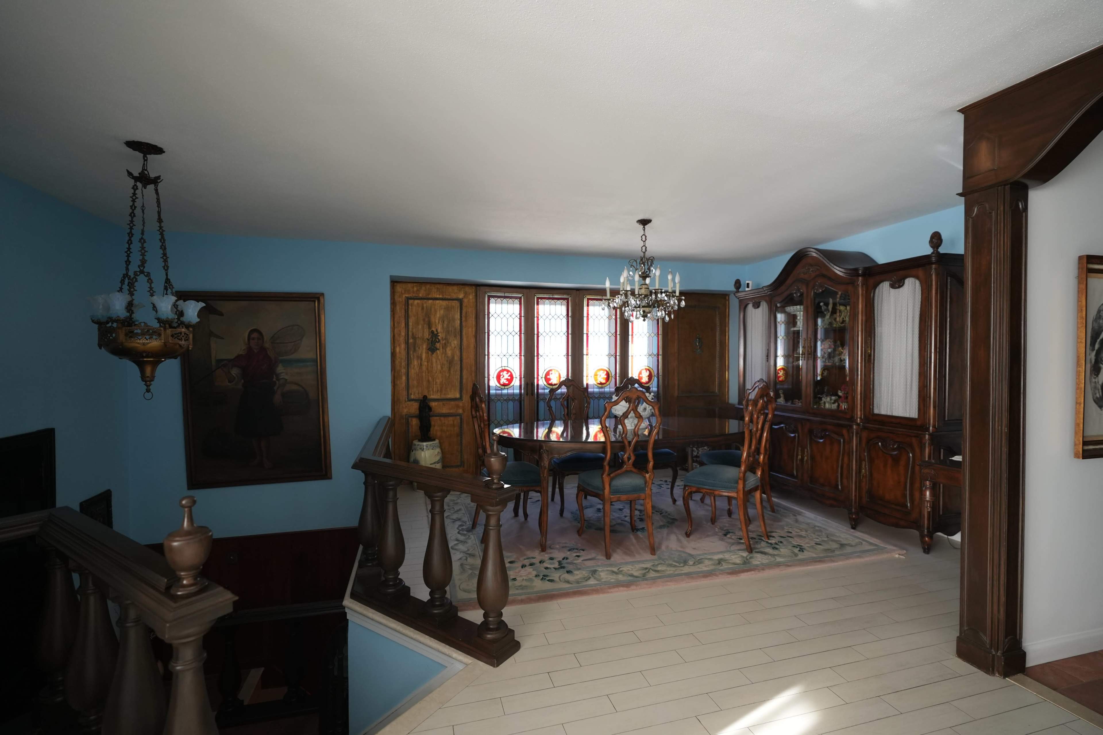 The image shows a well-furnished dining room featuring wooden furniture, a chandelier, and decorative stained glass windows.