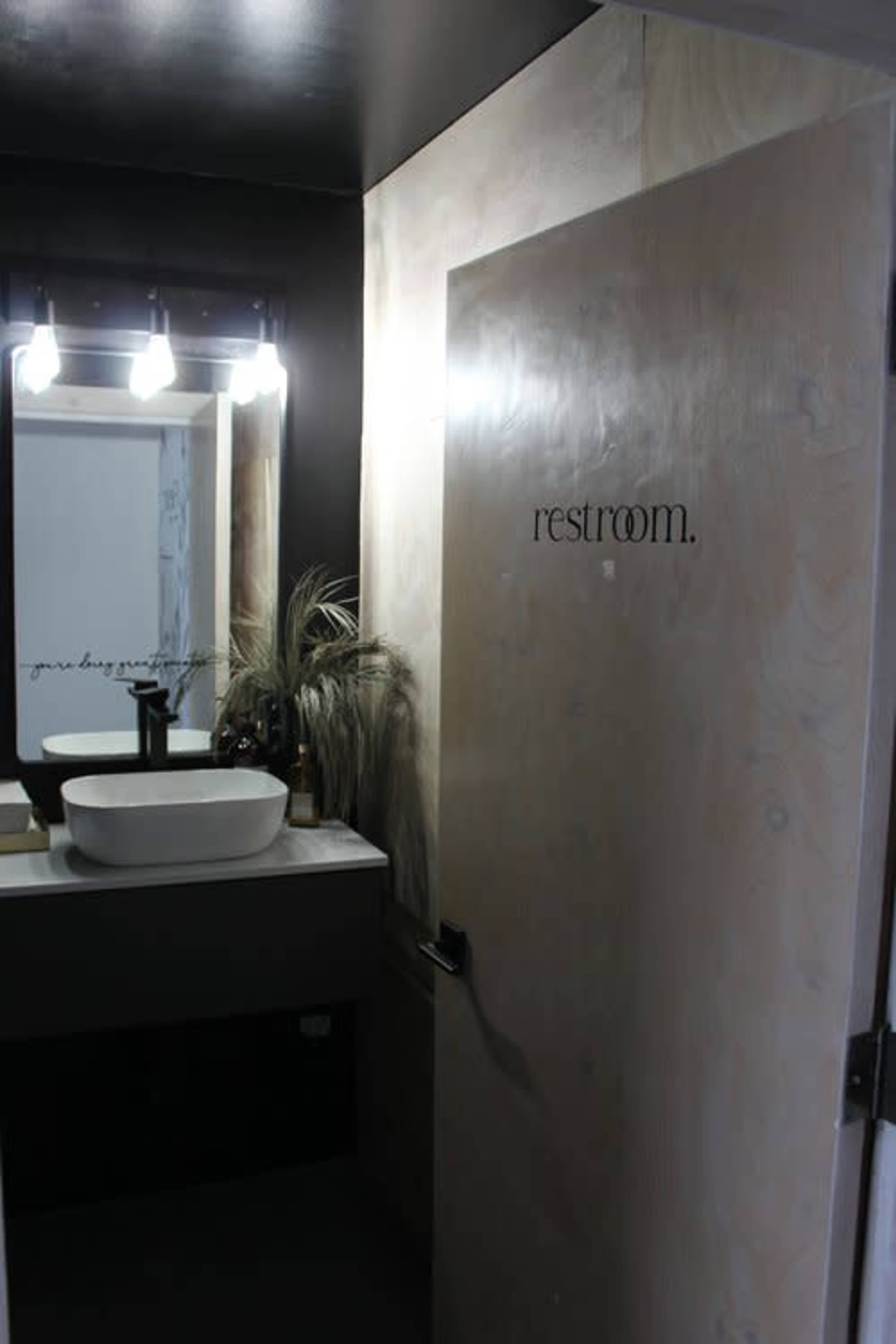 A modern restroom with a minimalist design featuring a wooden door labeled "restroom," a sink, and a mirror illuminated by overhead lights.