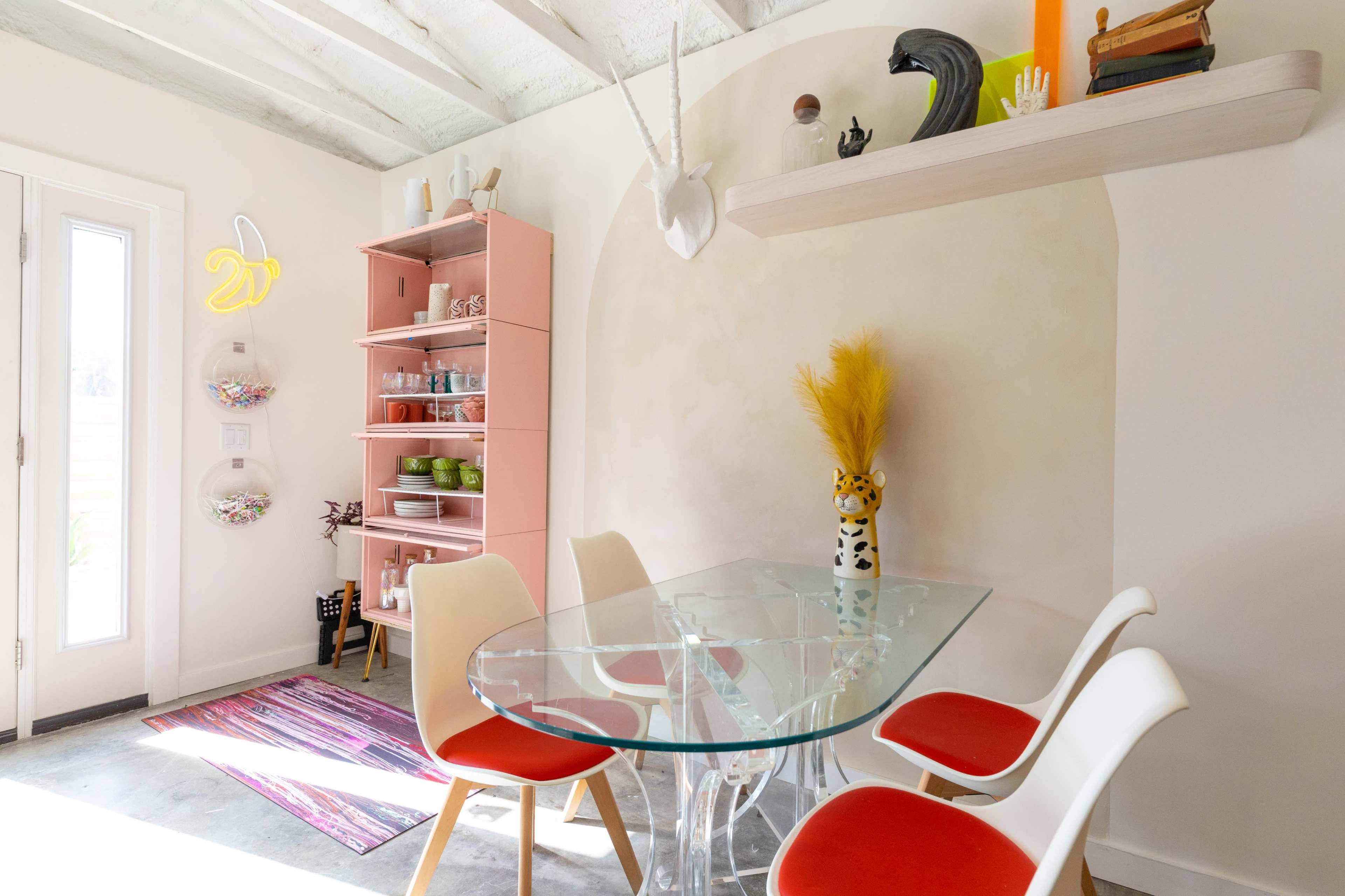 A glass dining table surrounded by white and red chairs is set against a pink shelving unit and a light-colored wall in a well-lit, minimalistic interior.