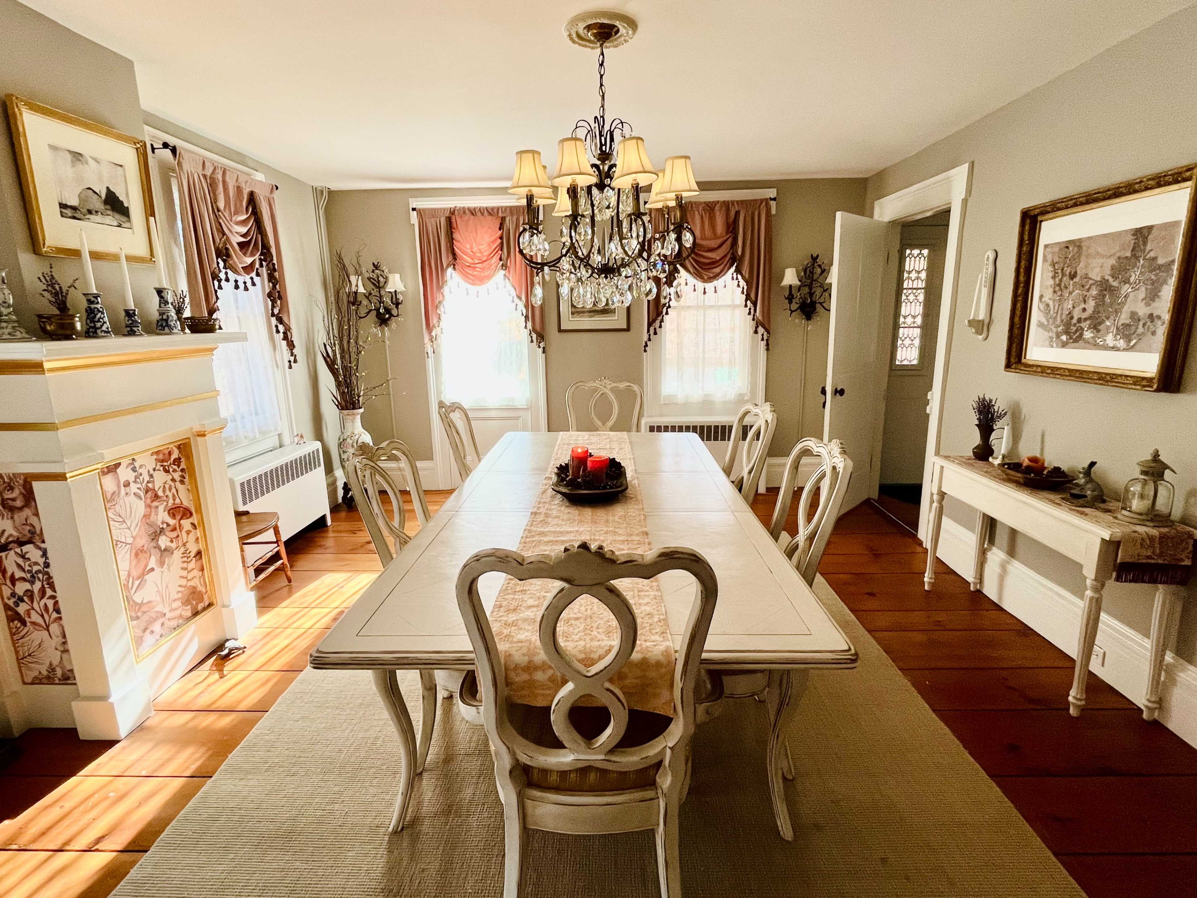 A well-lit dining room features a long table surrounded by ornate chairs, a chandelier overhead, and decor including curtains and wall art.