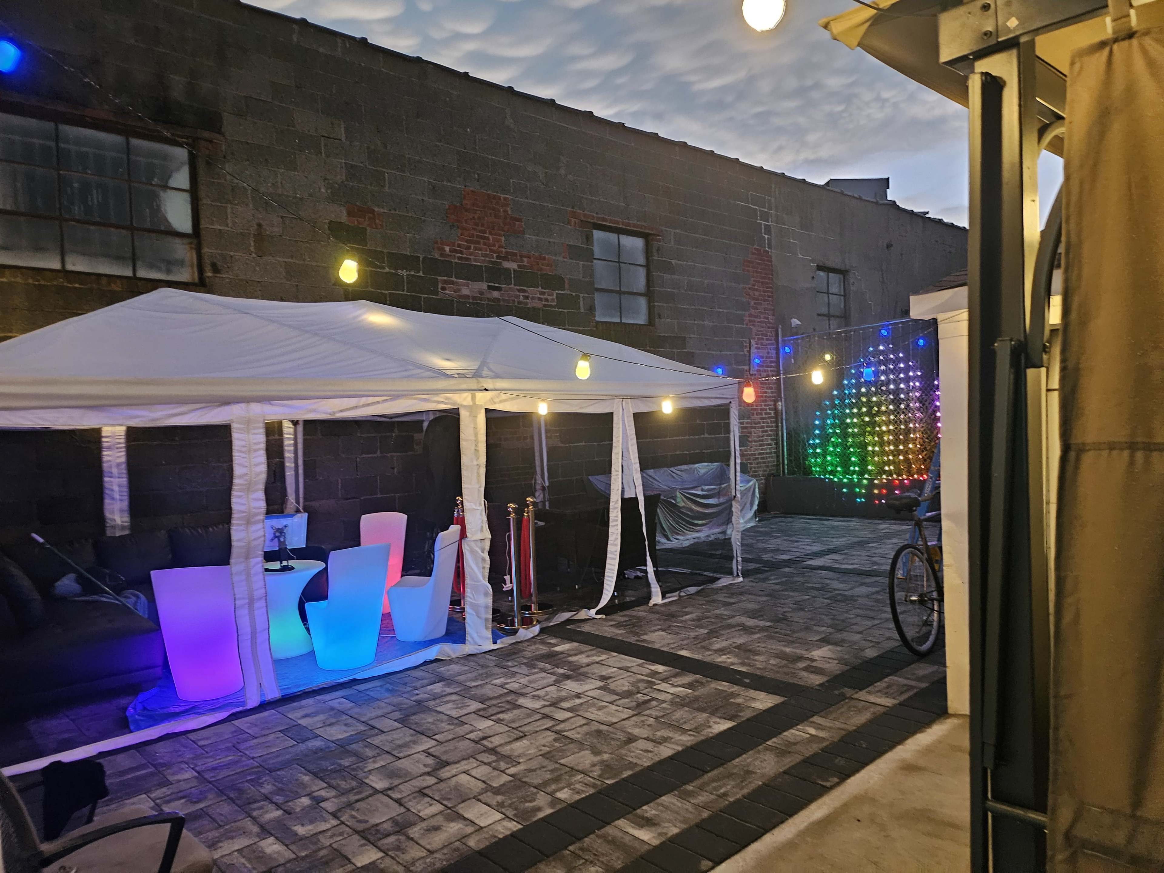 The image shows a patio area with a white tent, colorful LED lights, and a seating arrangement, against a backdrop of a brick building.
