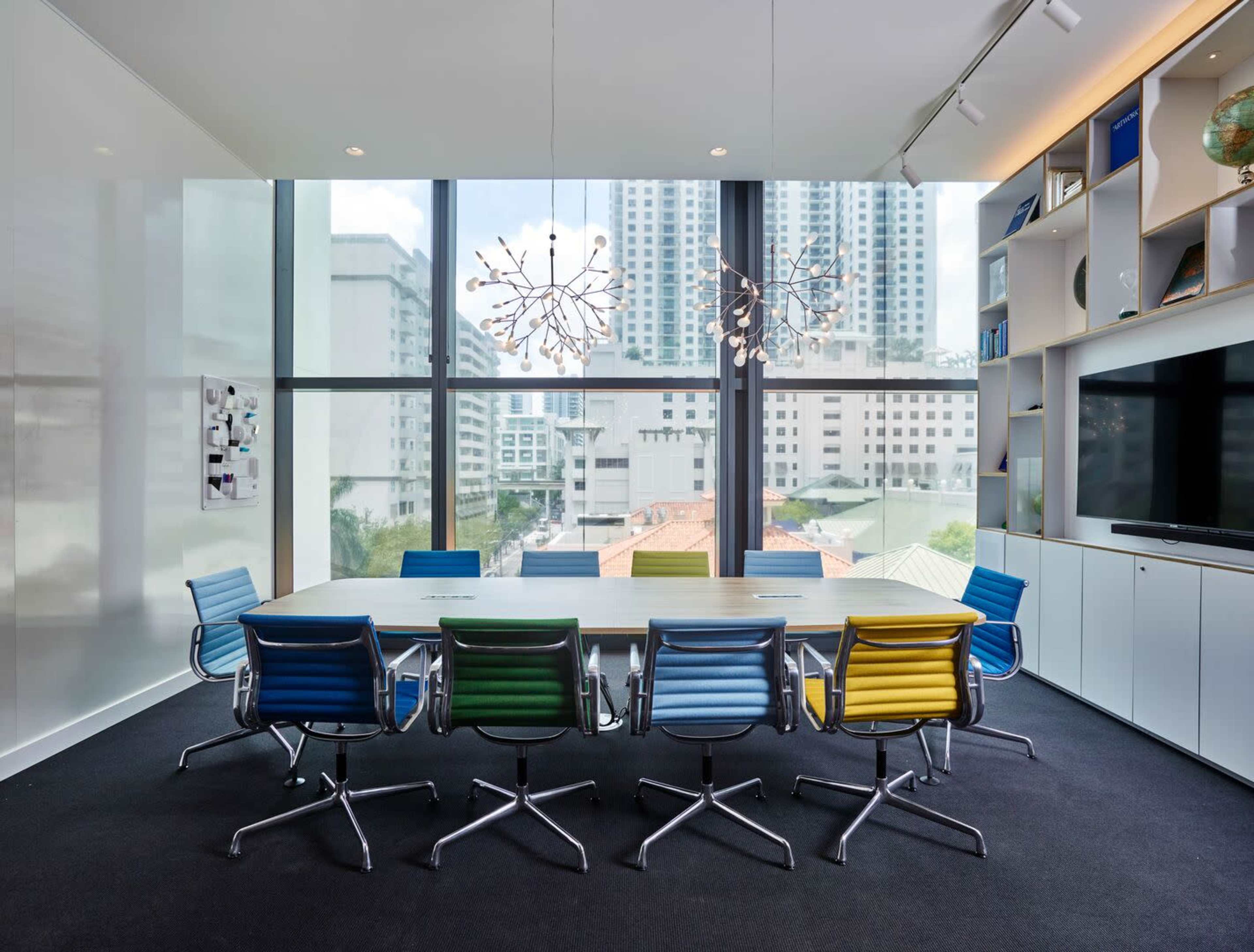 A modern conference room features a long table surrounded by colorful chairs and large windows showcasing a city view.