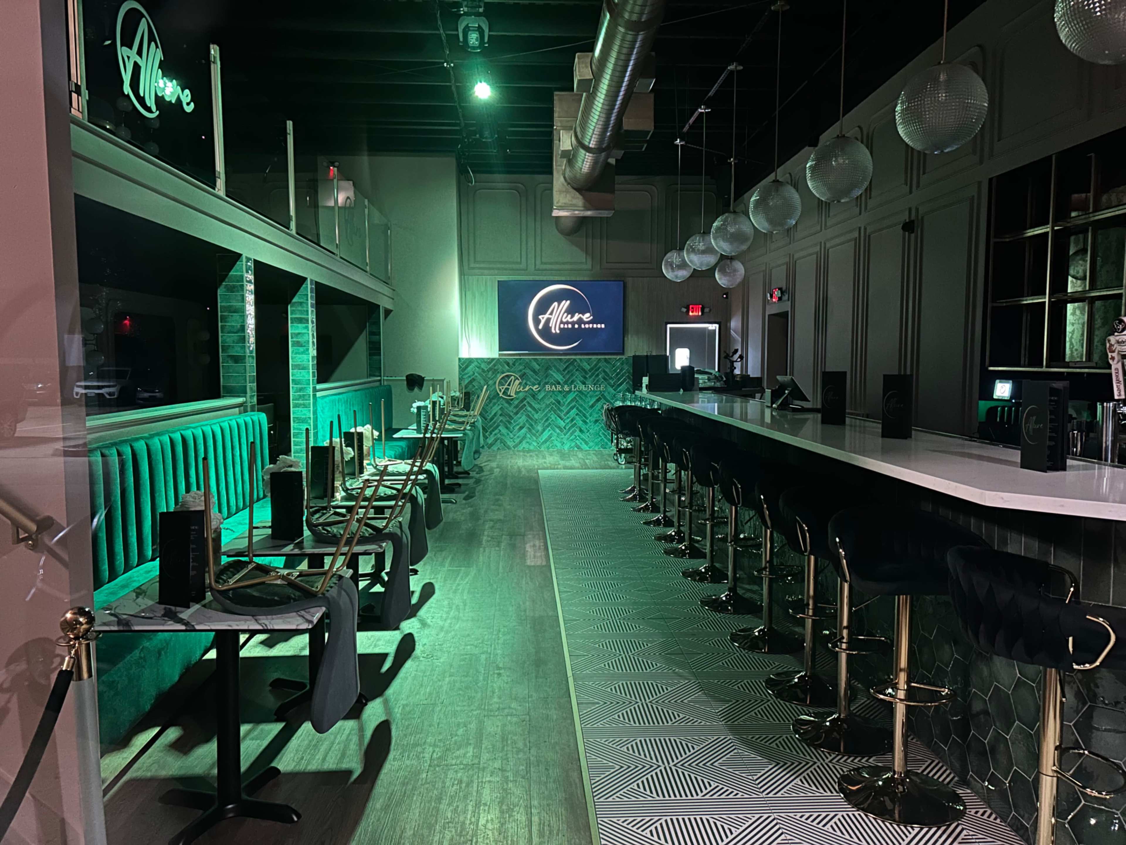 The image shows a modern bar interior with green lighting, featuring a long white countertop and stylish seating arrangements.