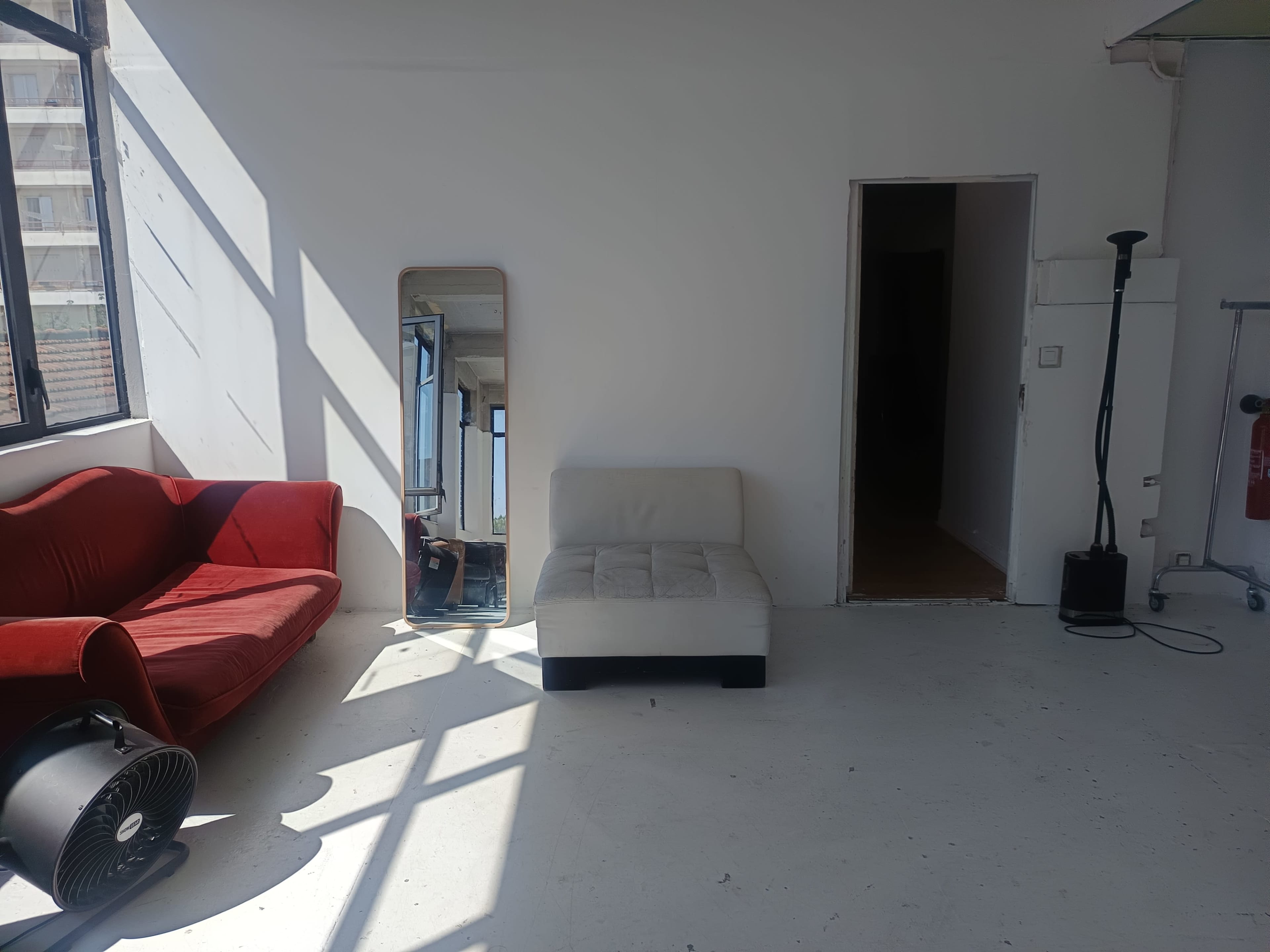 The image shows a sunlit room with a red couch, a white chair, a mirror, and a doorway leading to another space.