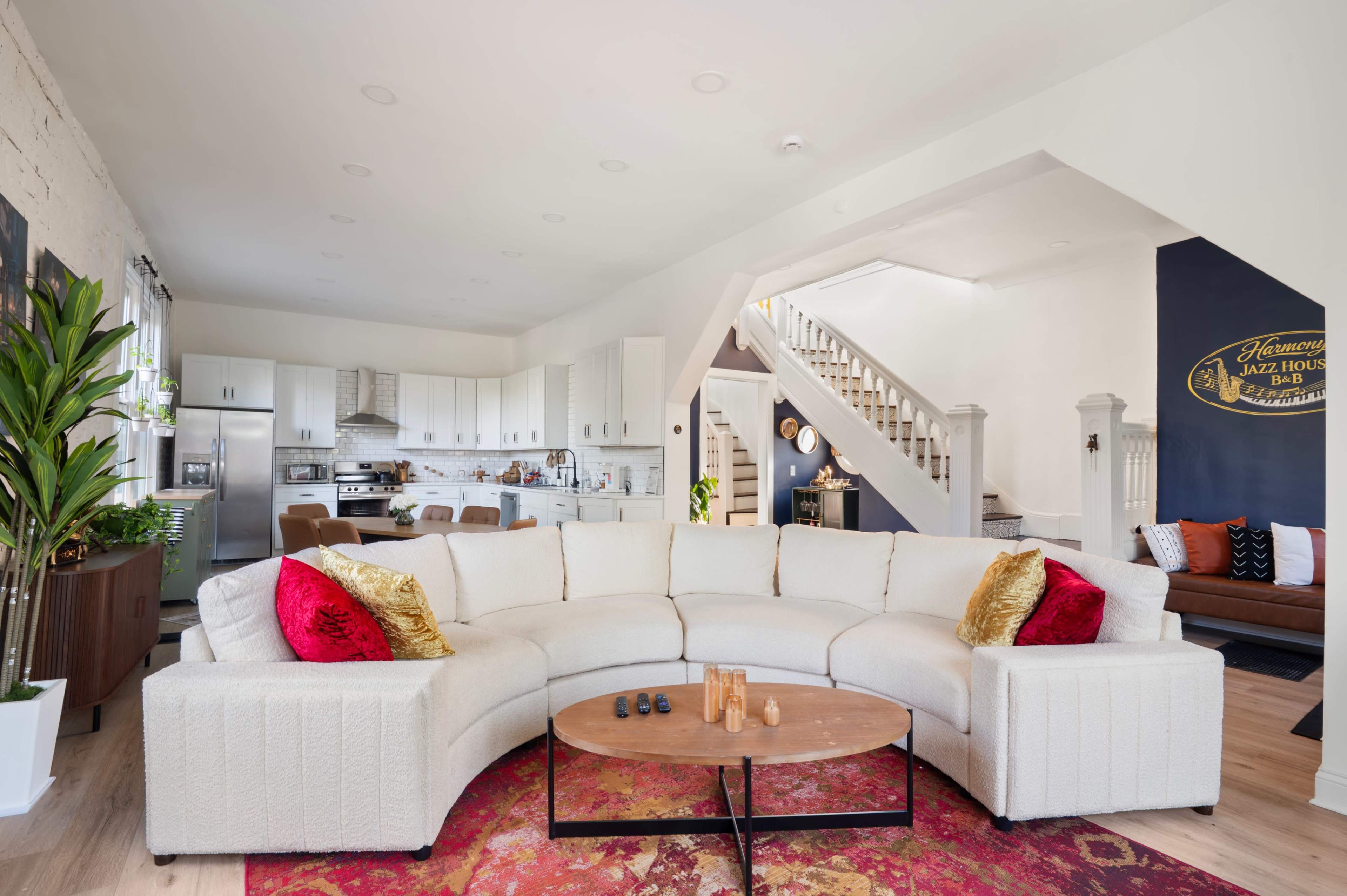 The image shows a modern living room with a curved white sectional sofa, a round coffee table, and a kitchen visible in the background.