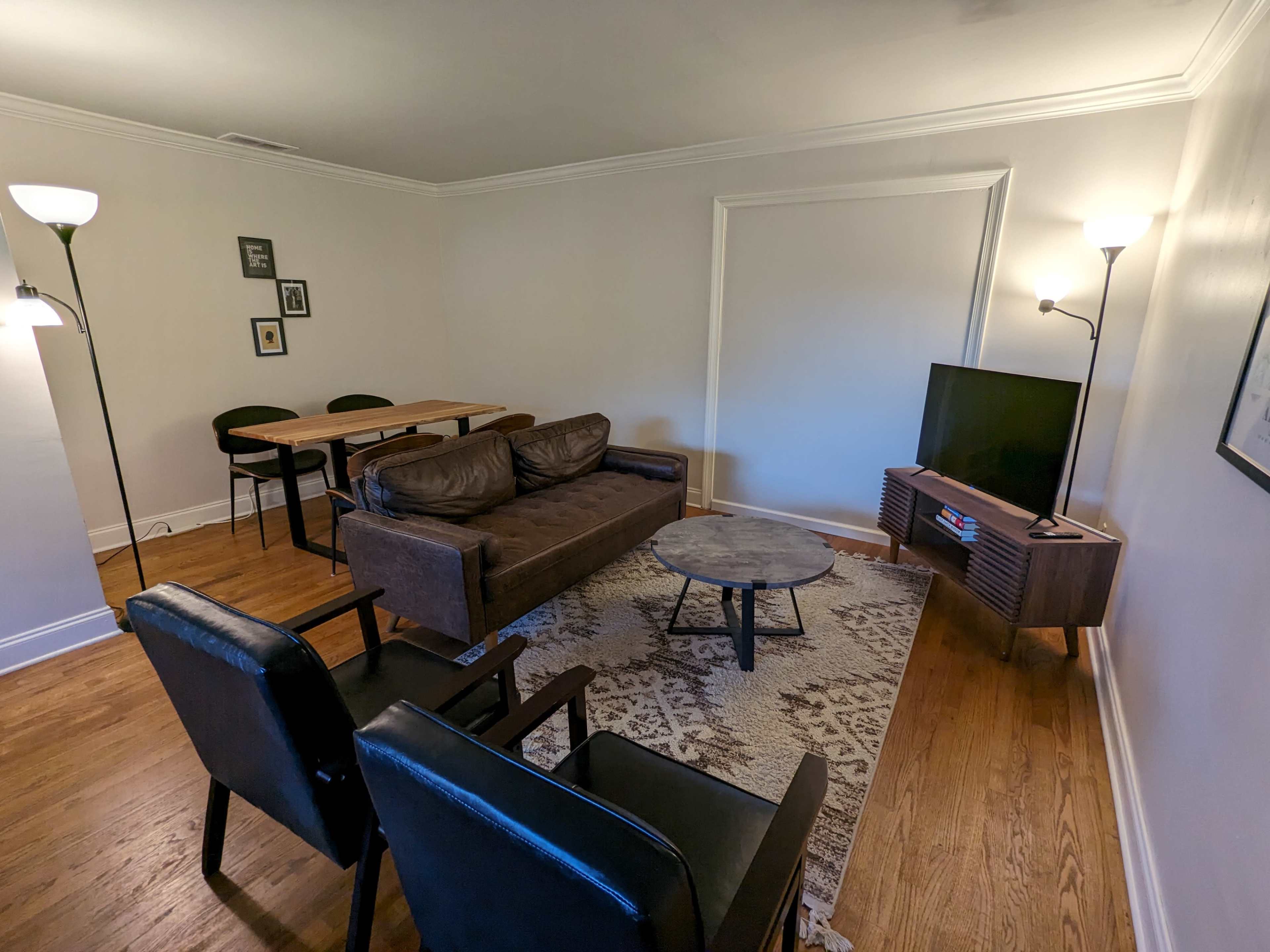 A living room features a brown sofa, two black chairs, a round table, a TV on a stand, and a patterned area rug on hardwood flooring.