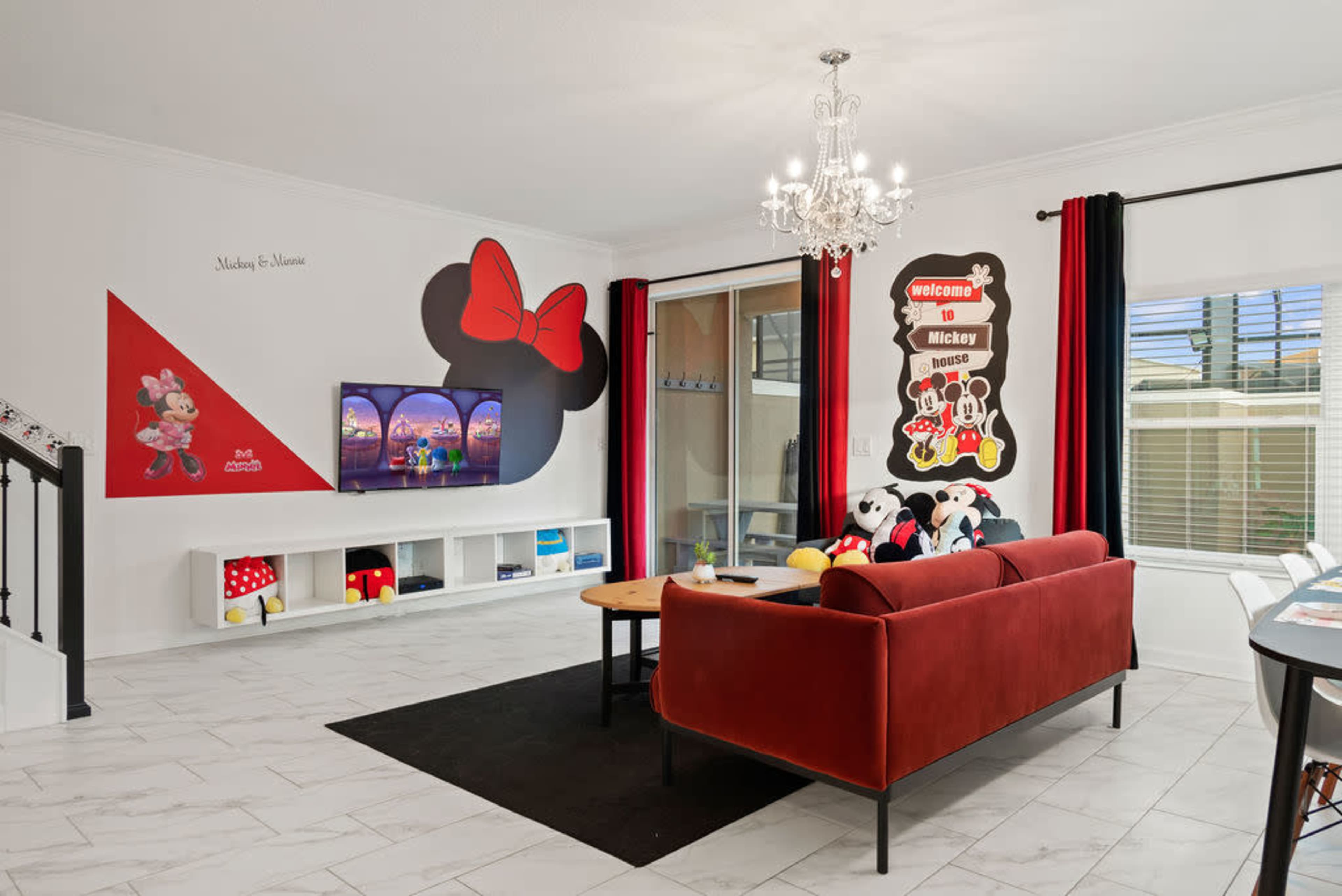 A living room decorated with Mickey Mouse-themed artwork, featuring a red couch, a large television, and playful accents like stuffed toys and colorful decor.