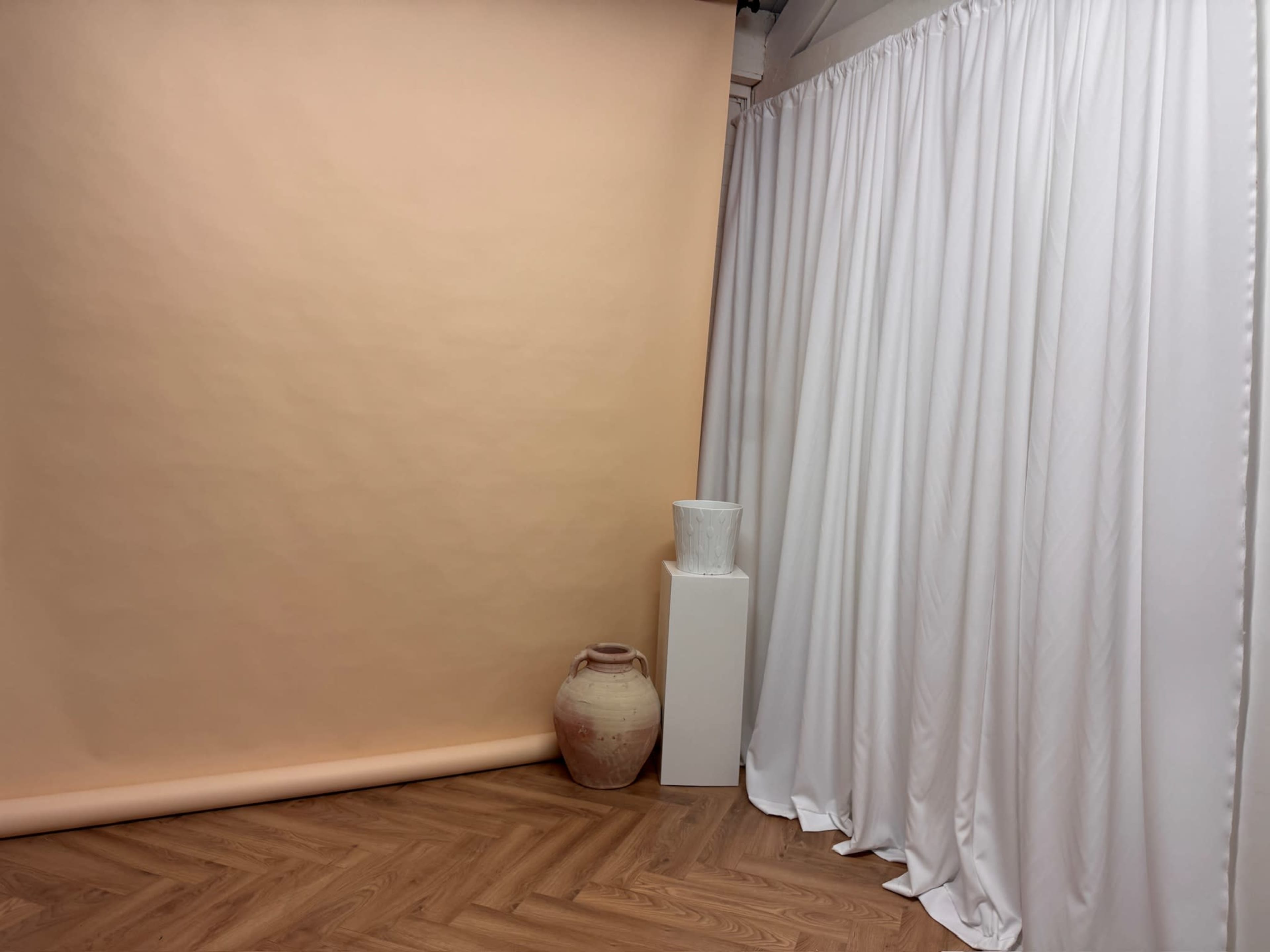 The image shows a corner of a studio with a peach-colored backdrop, white curtains, a white pedestal with a decorative vase, and a larger pottery jar on the floor.