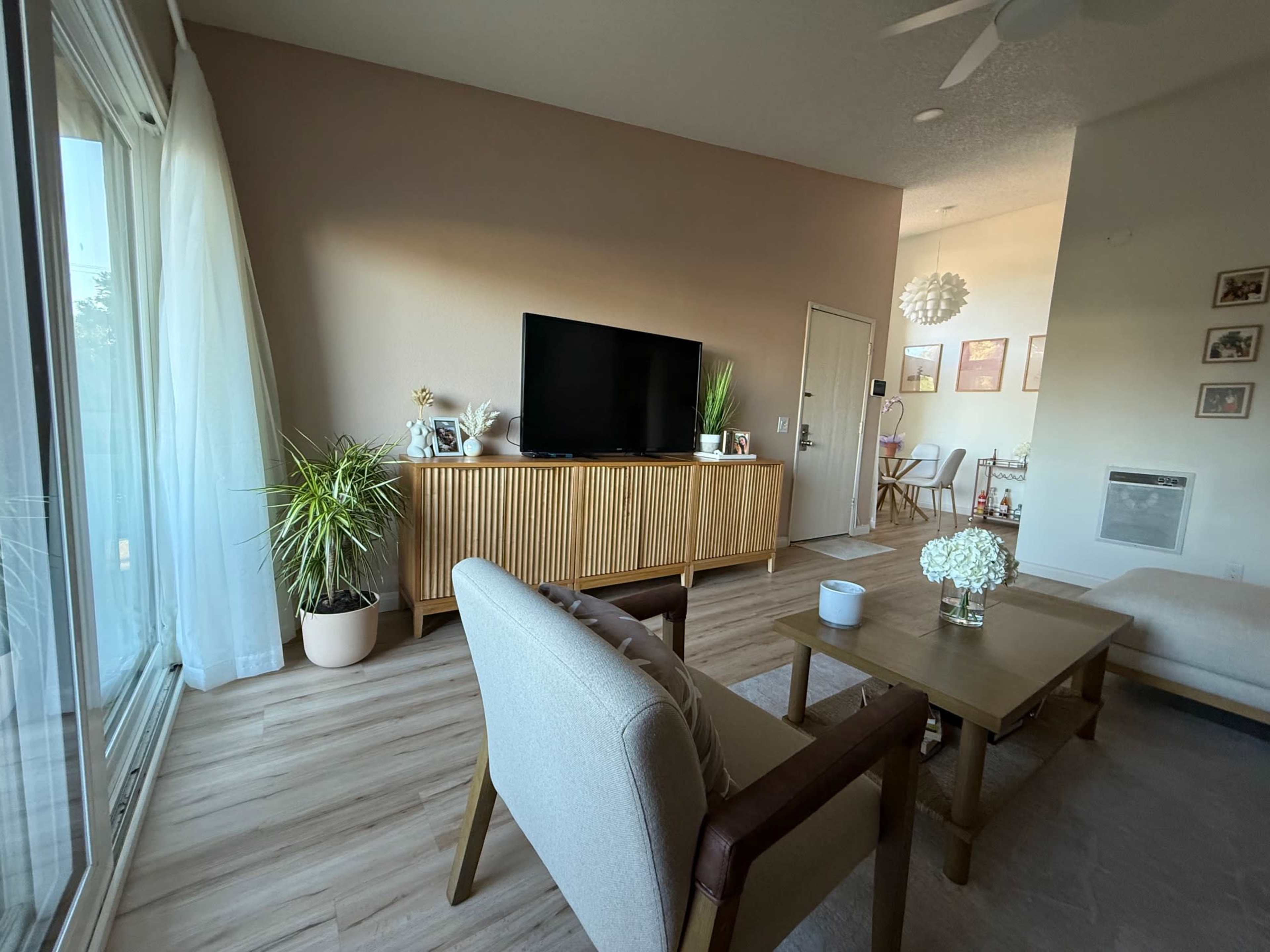 The image shows a modern living room with a wooden TV stand, a coffee table, and a comfortable seating area, illuminated by natural light from large windows.