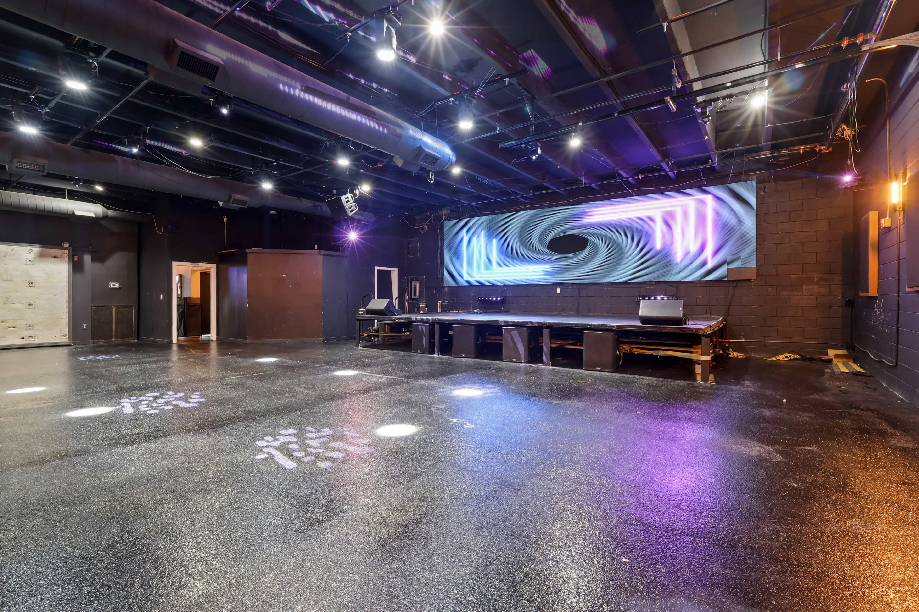 The image shows a large, empty performance space with a glossy floor, colorful lighting, and a large screen displaying abstract visuals on the far wall.