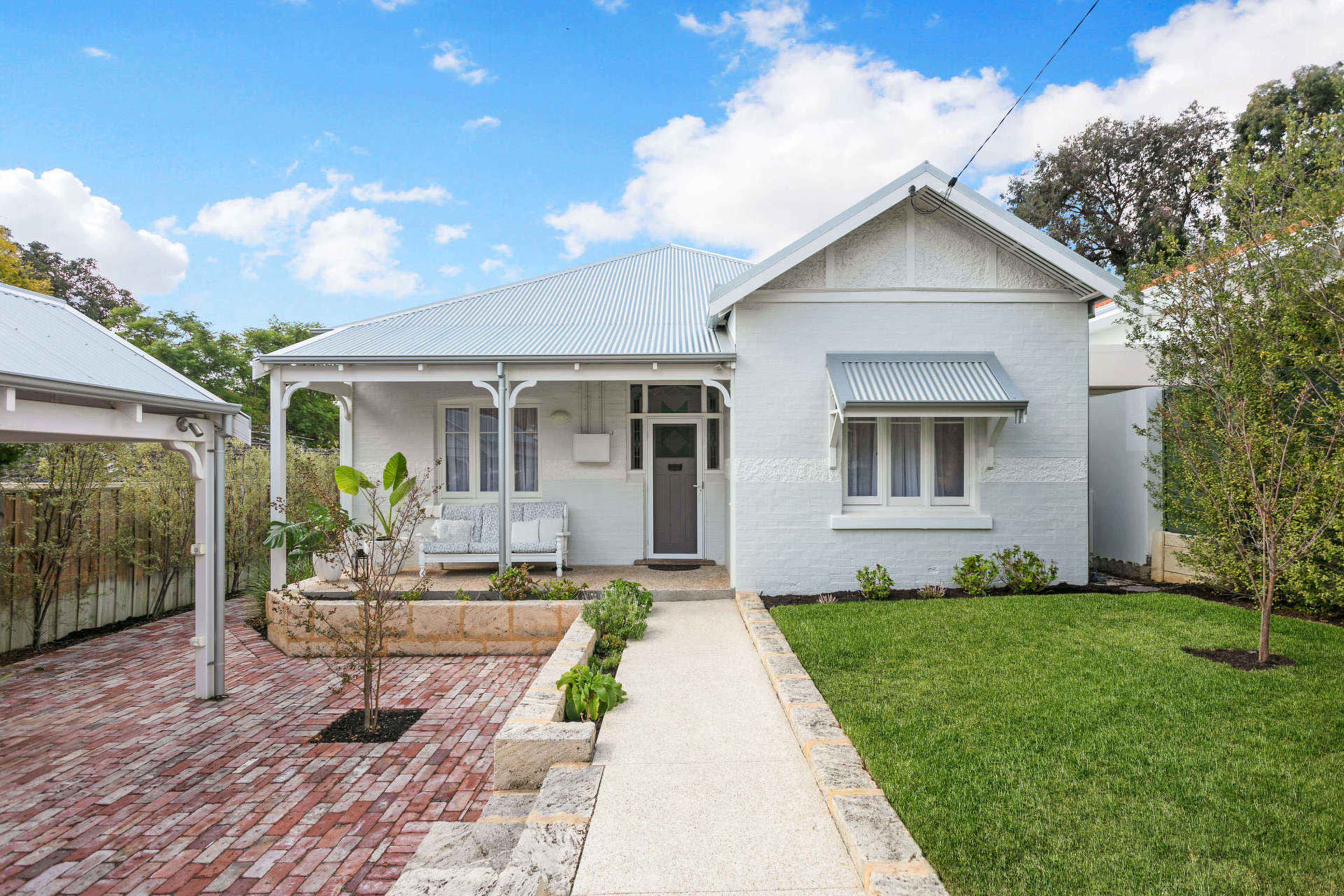 A white cottage with a light blue tin roof, featuring a front porch and a landscaped yard with brick pathways and greenery.
