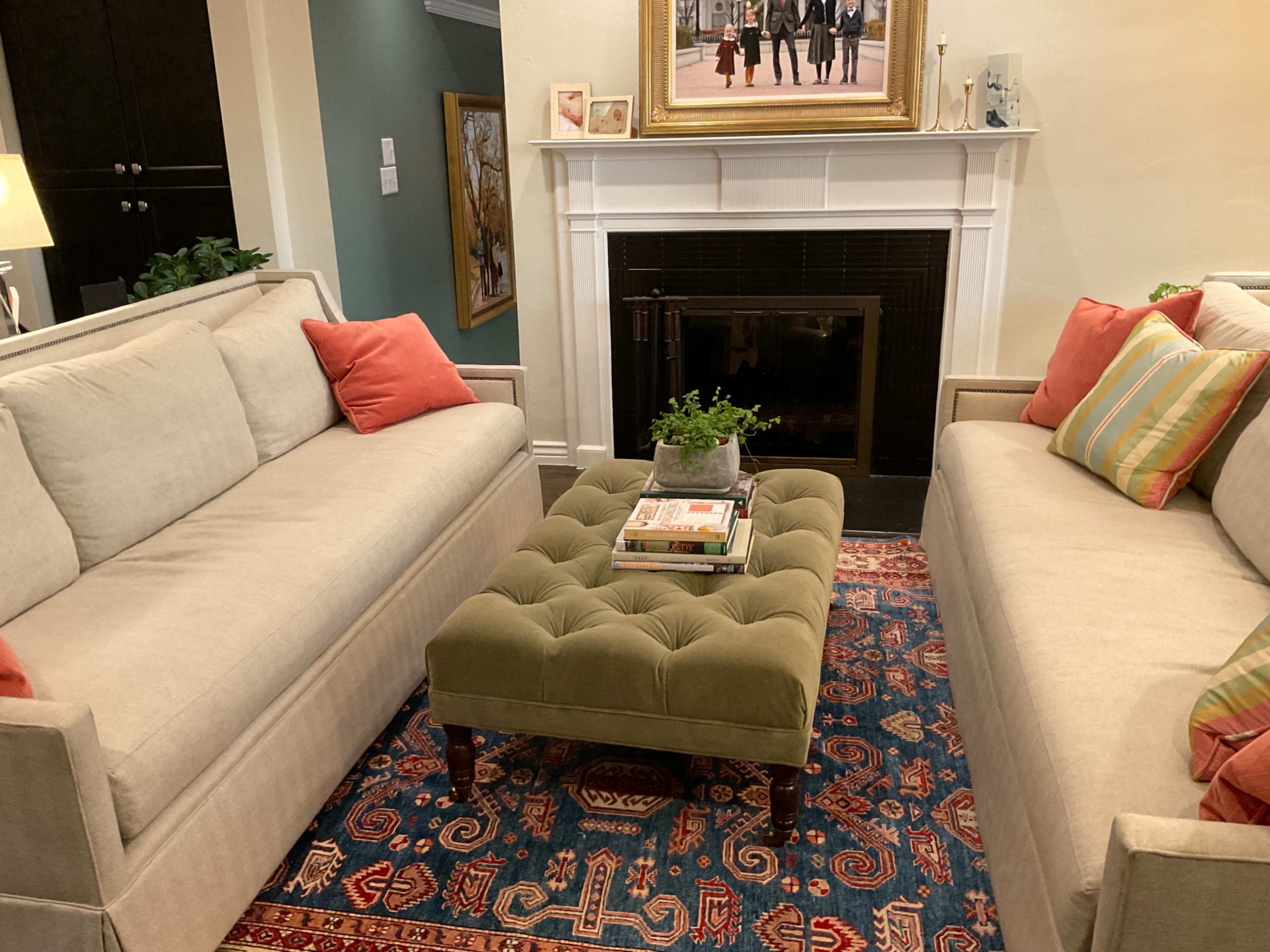 A living room with two beige sofas facing each other, a green tufted coffee table in the center, and a fireplace with a decorative frame and artwork above it.