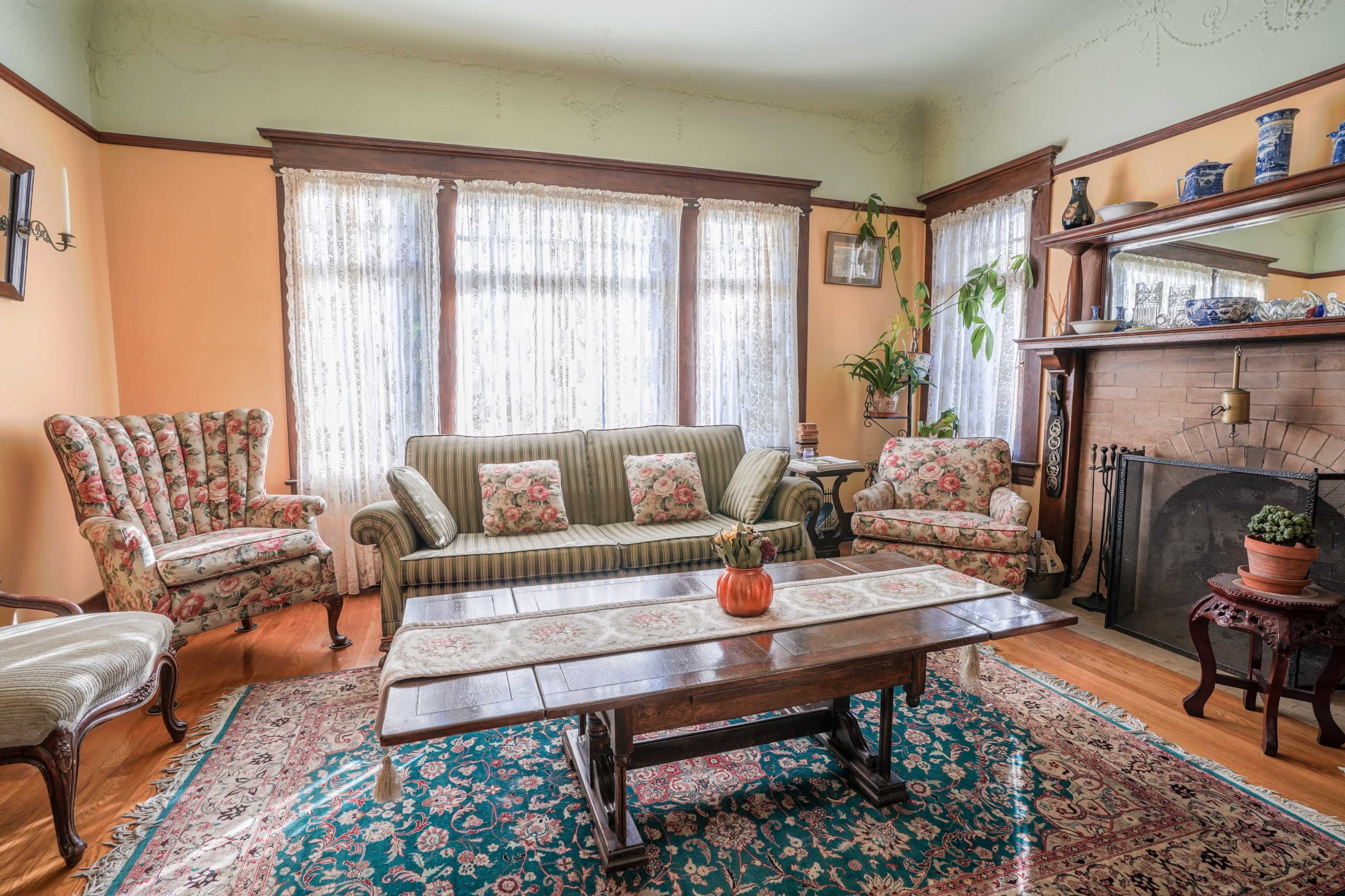 A cozy living room with a floral-patterned sofa, matching armchairs, a wooden coffee table, and a brick fireplace, all surrounded by large windows with lace curtains.