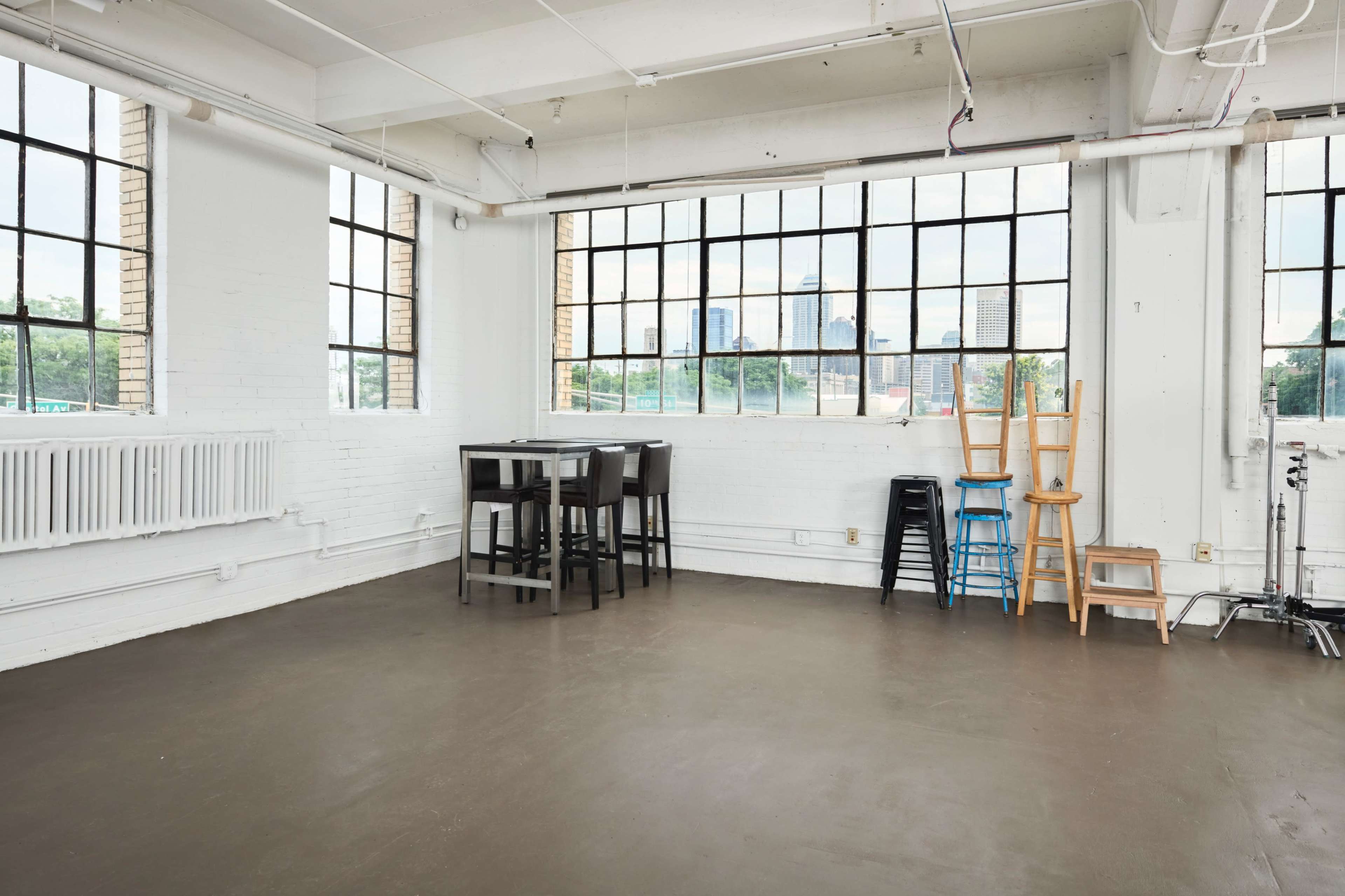The image shows a spacious, empty studio with large windows overlooking a city skyline, furnished with a table and chairs.