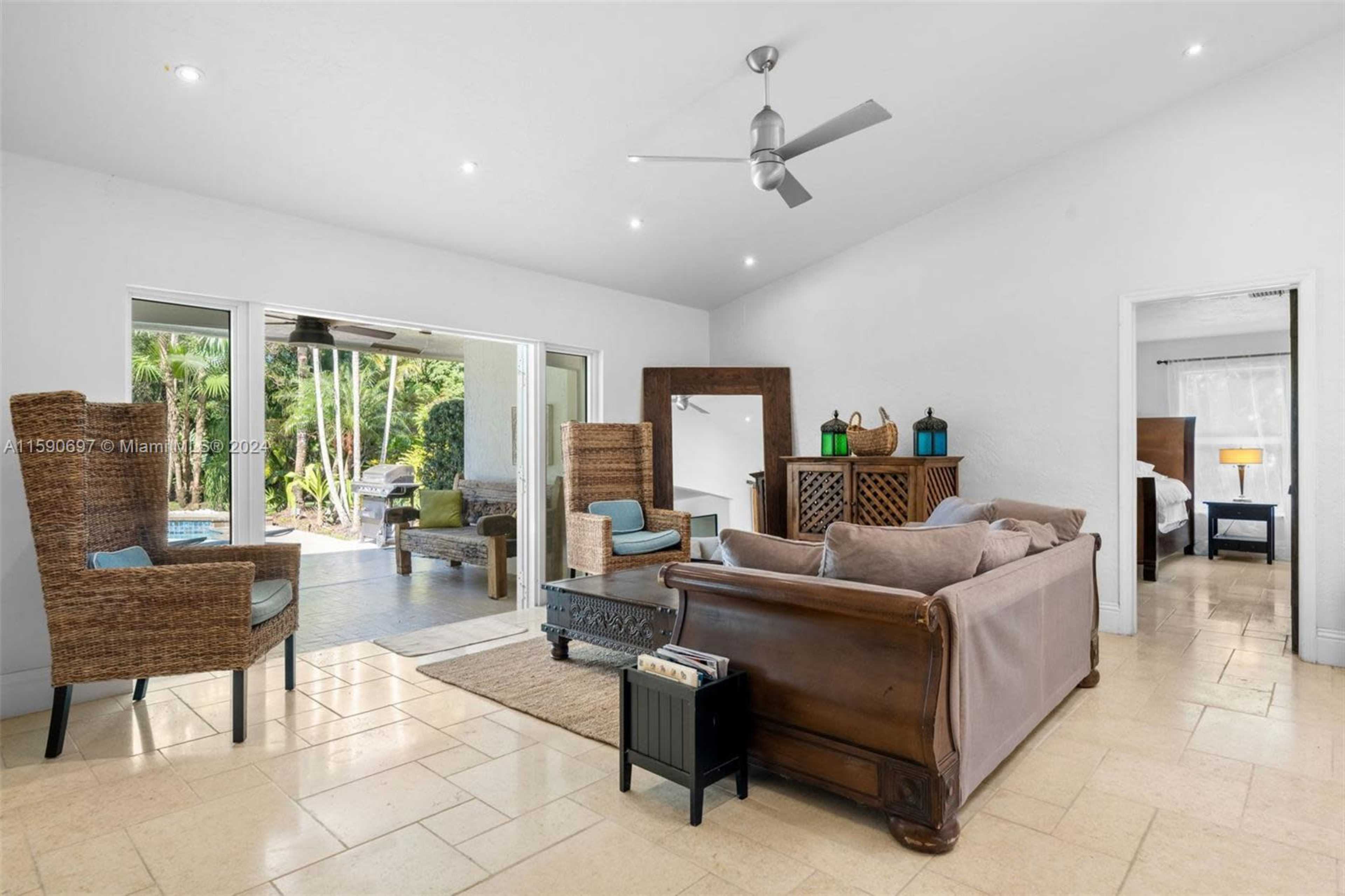 A spacious living room features a brown sofa, two woven armchairs, and a coffee table, with large windows providing views of an outdoor patio and garden.