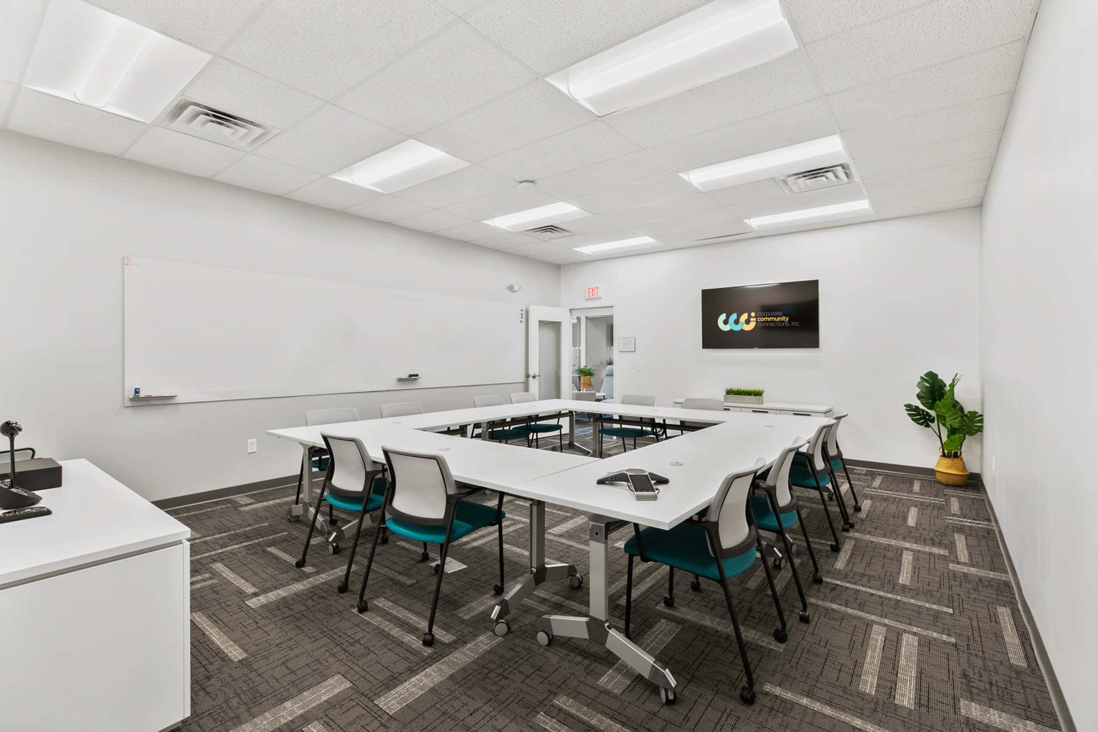 A conference room is set up with a rectangular arrangement of white tables and chairs, featuring a whiteboard, a television screen, and a small plant in the corner.