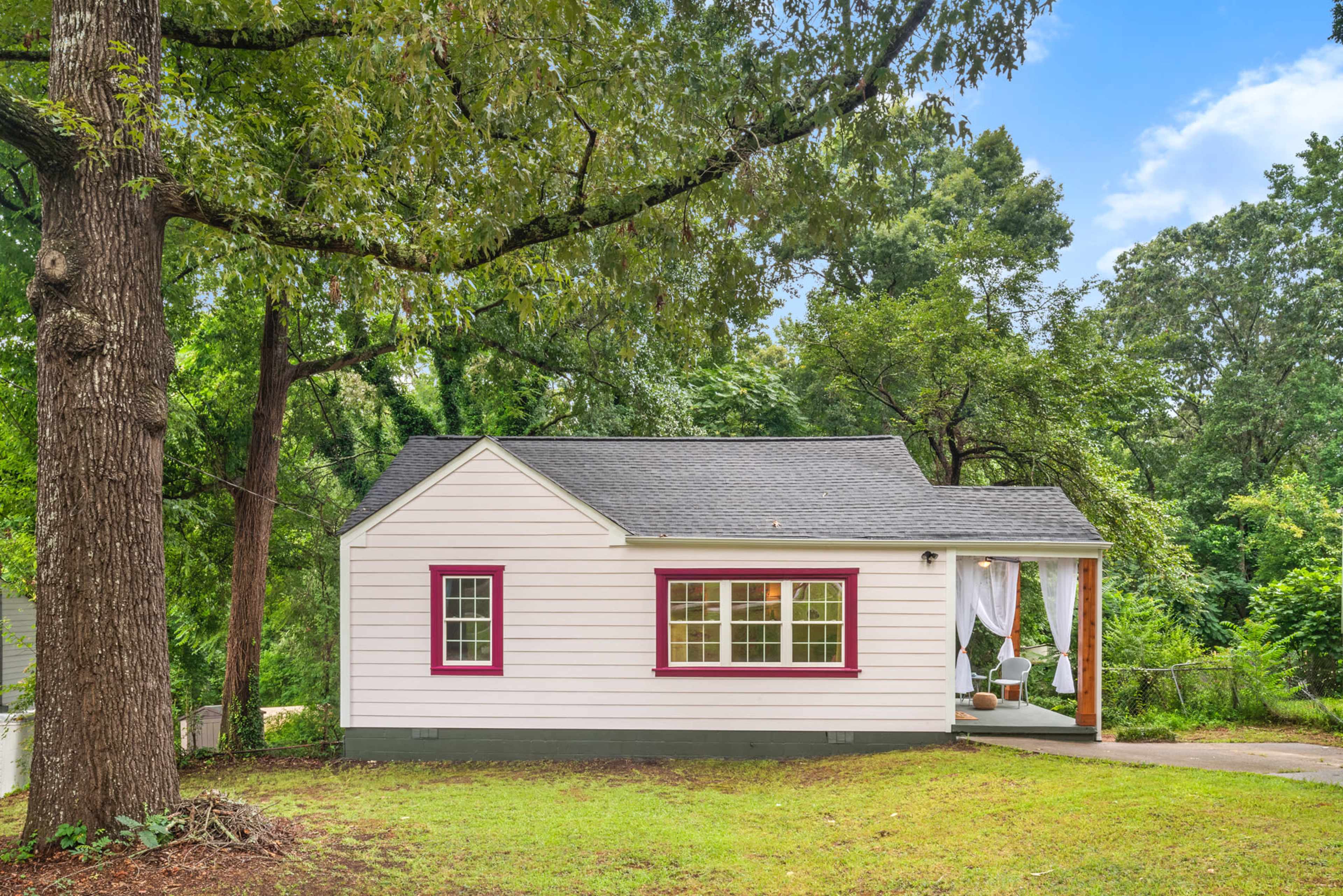 A one-story house with a white exterior, red trim, and a covered porch is surrounded by trees and a grassy area.