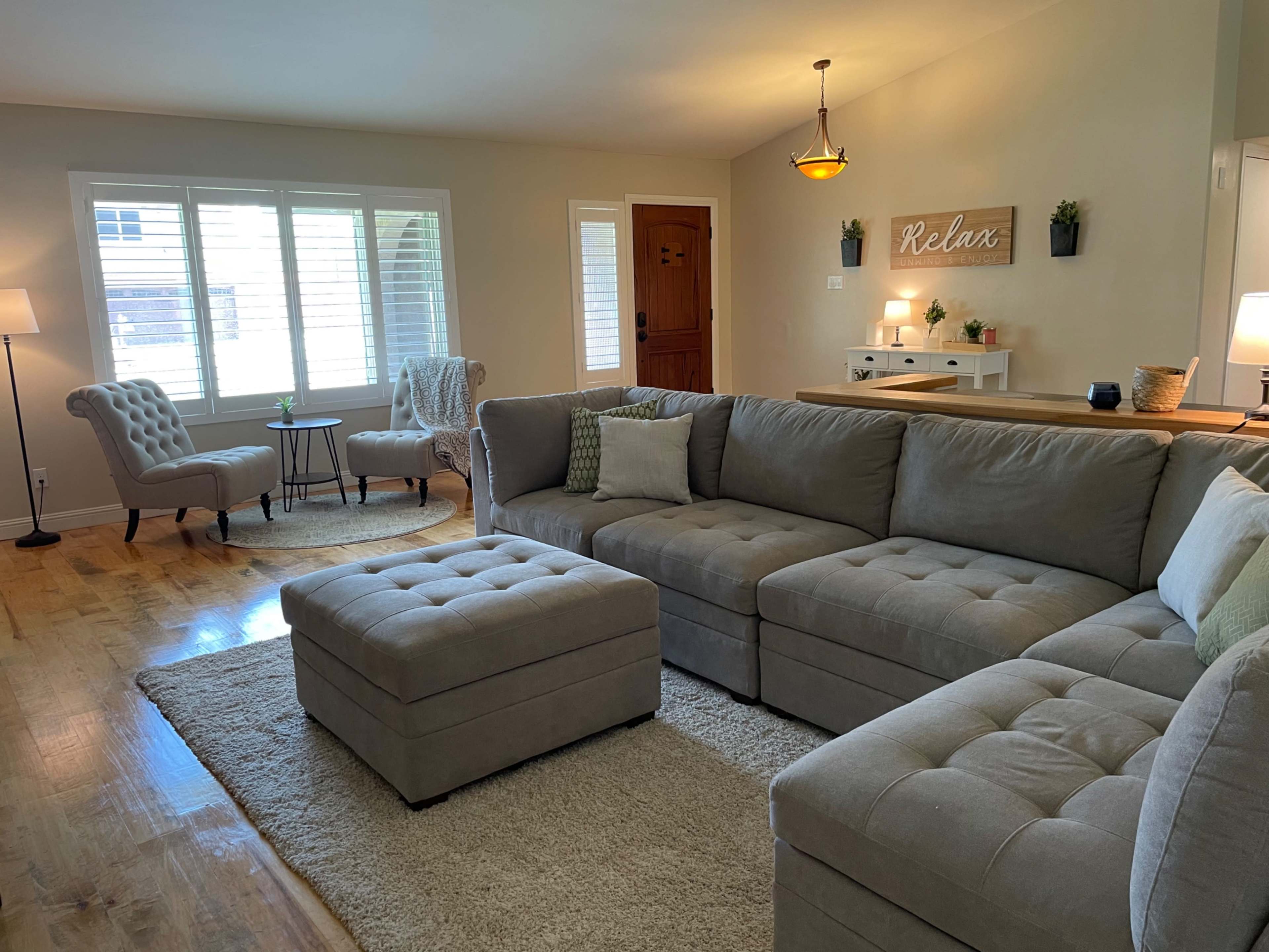 A light-filled living room features a gray sectional sofa, an ottoman, two armchairs, and a wooden door.