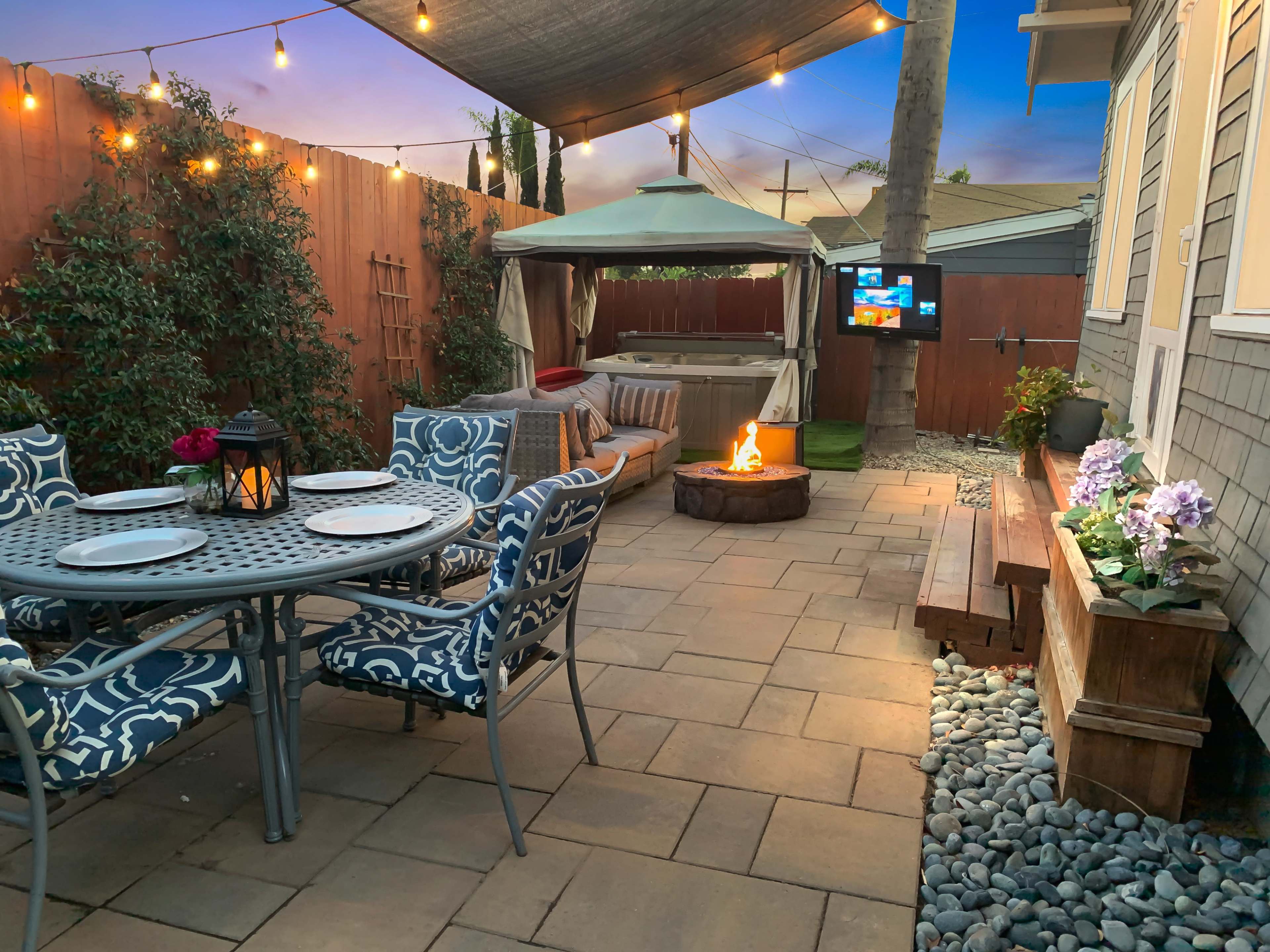 The image shows a backyard patio with a dining table, a fire pit, a seating area under a canopy, and a television mounted on the wall.