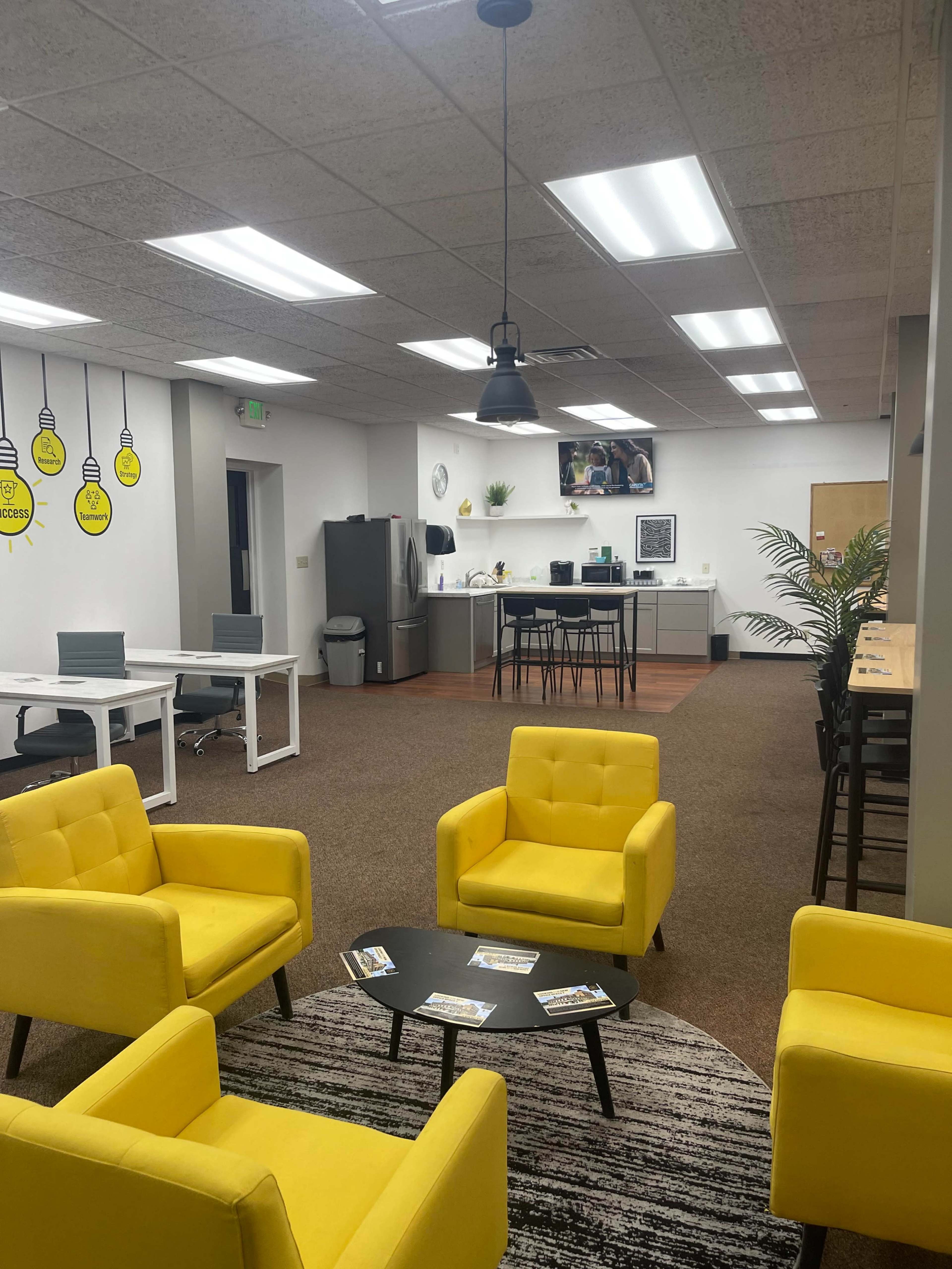 The image shows a modern office space with yellow seating, a kitchen area in the background, and several workstations.
