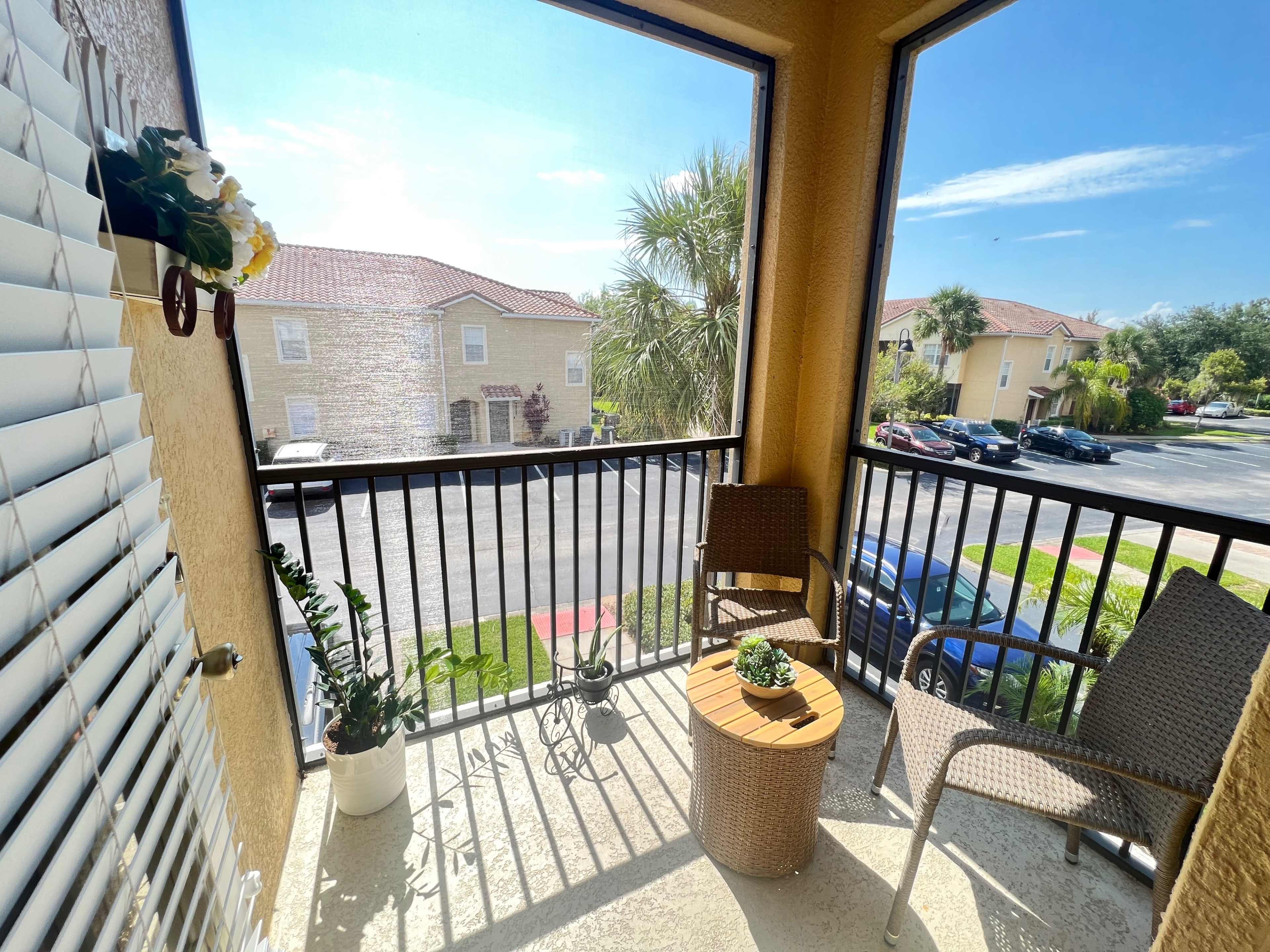 The image shows a small balcony with two chairs, a round table, and potted plants, overlooking a parking area and nearby buildings.