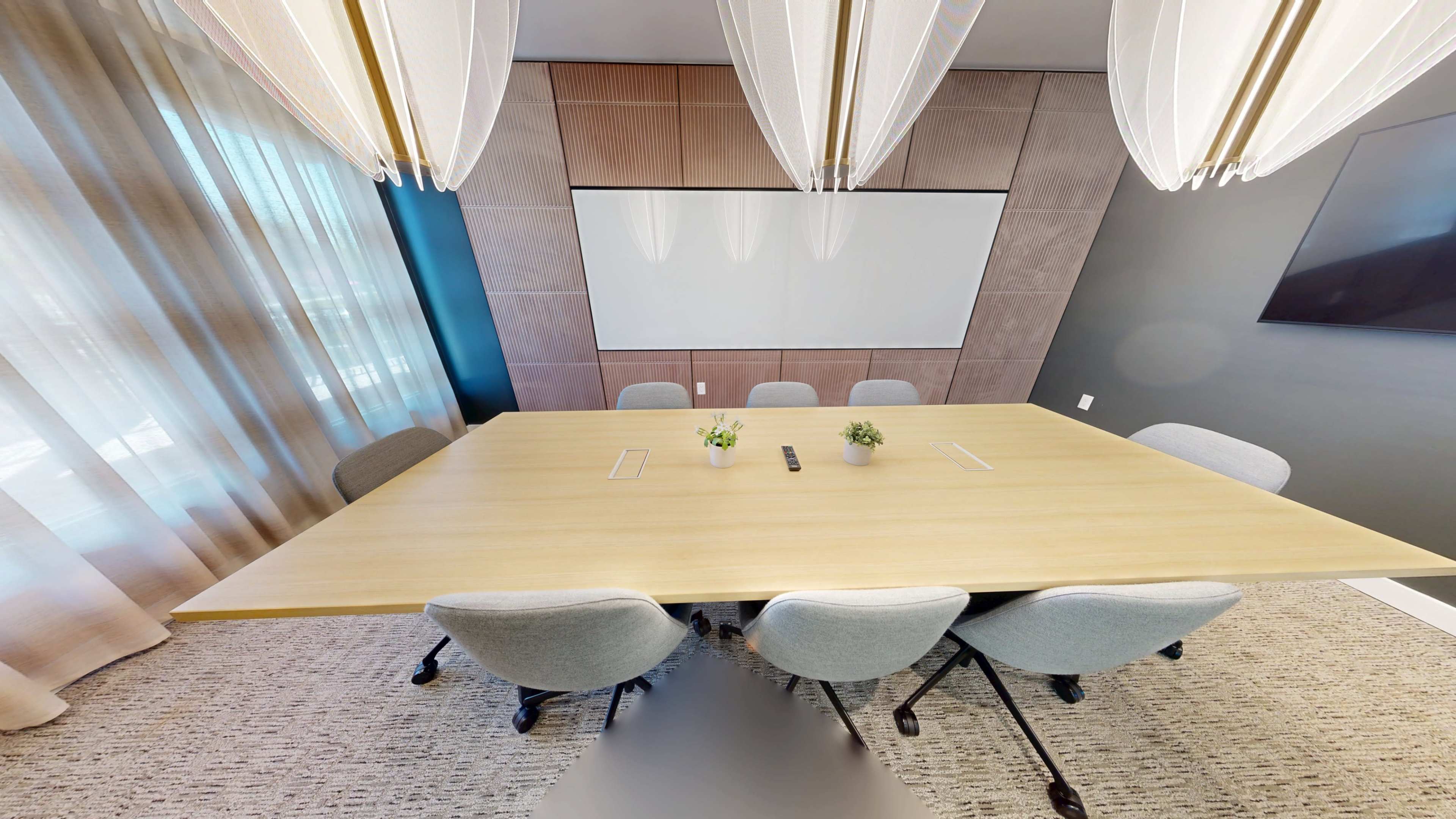 A modern conference room features a large rectangular wooden table surrounded by gray chairs, with a whiteboard and a television mounted on the walls.