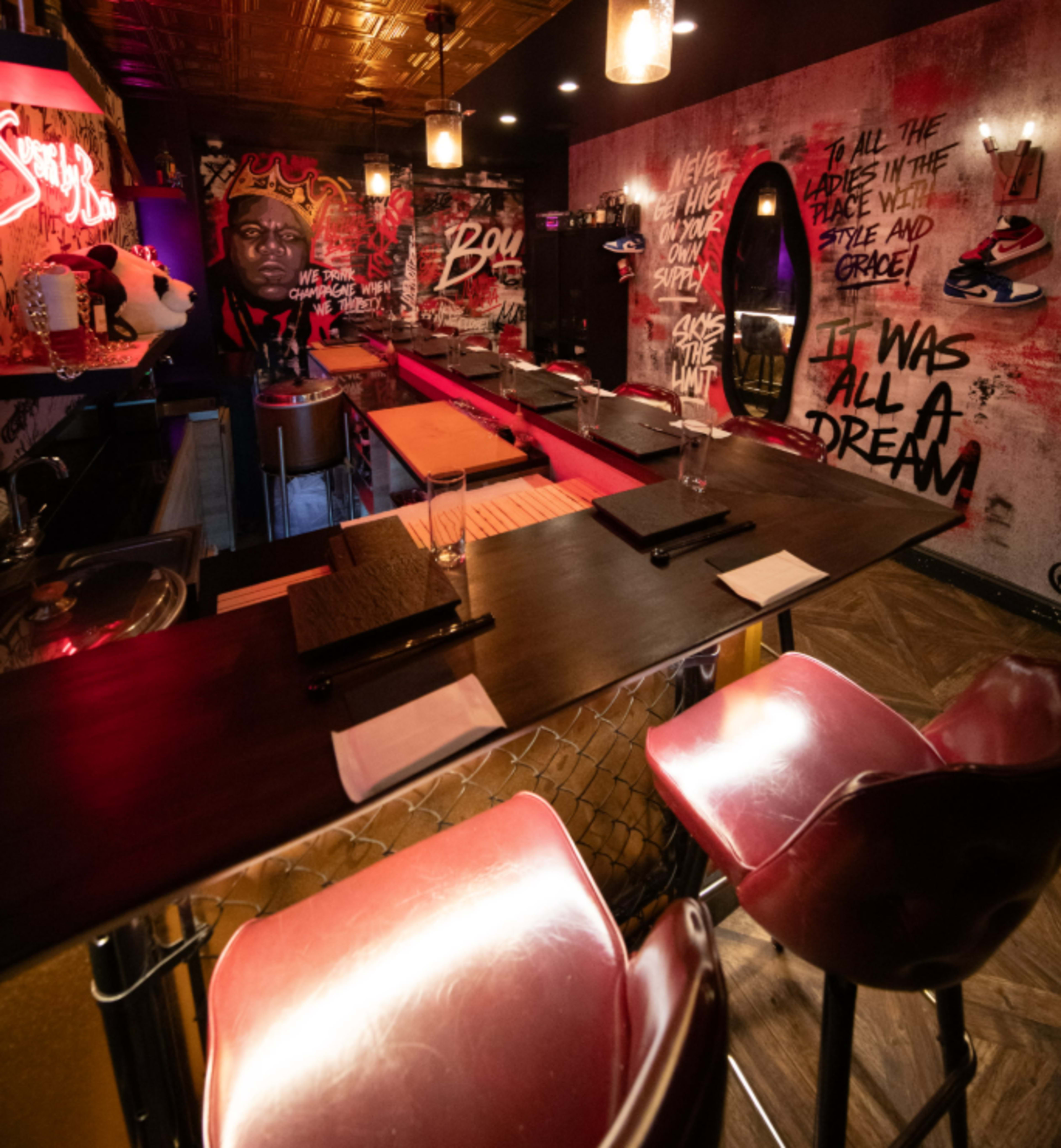 The image displays a dimly lit bar with red and black decor, featuring a long countertop with empty glasses and several barstools, adorned with graffiti and wall art.