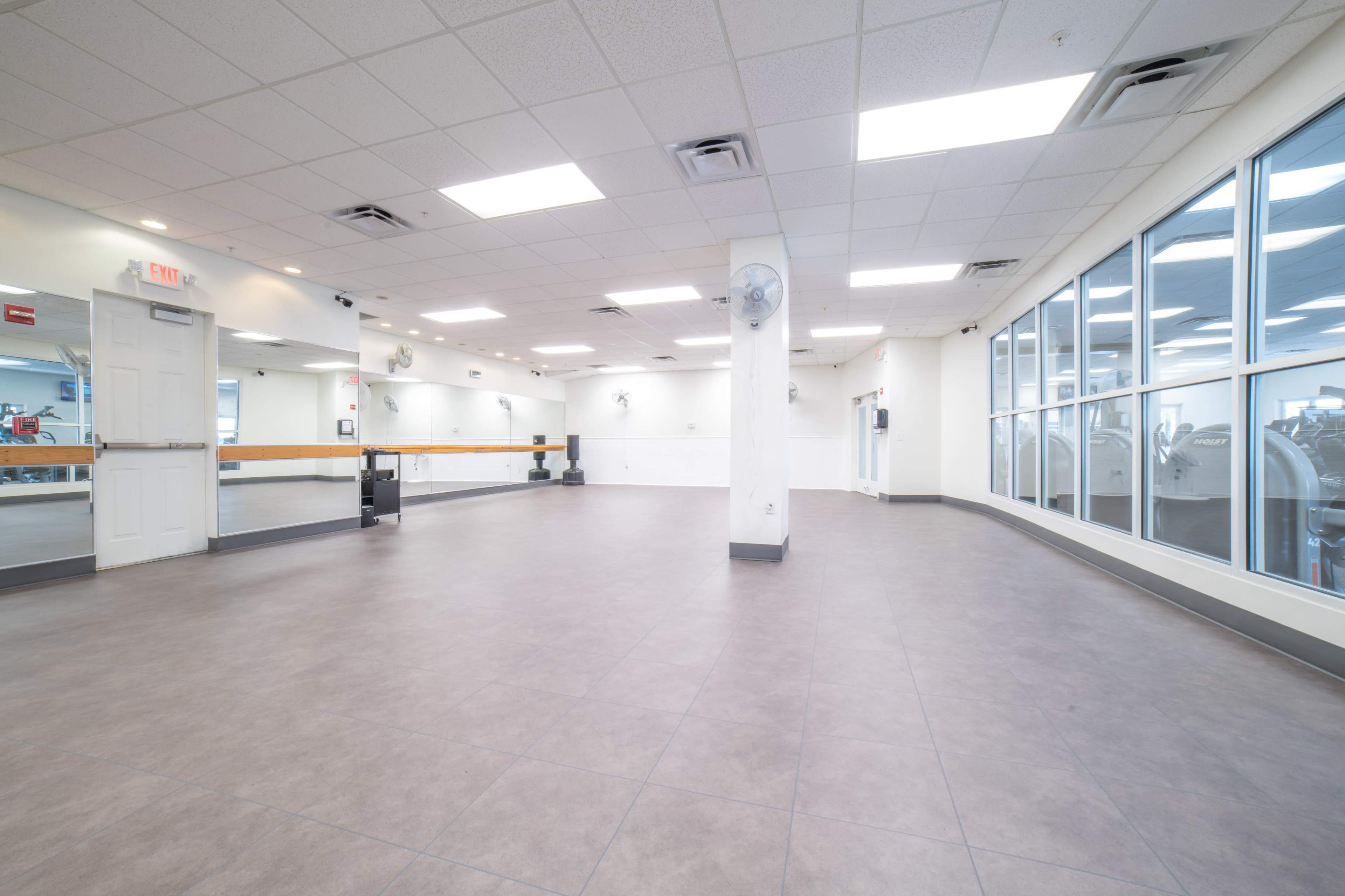 The image shows an empty fitness studio with mirrored walls and large windows allowing natural light to enter.