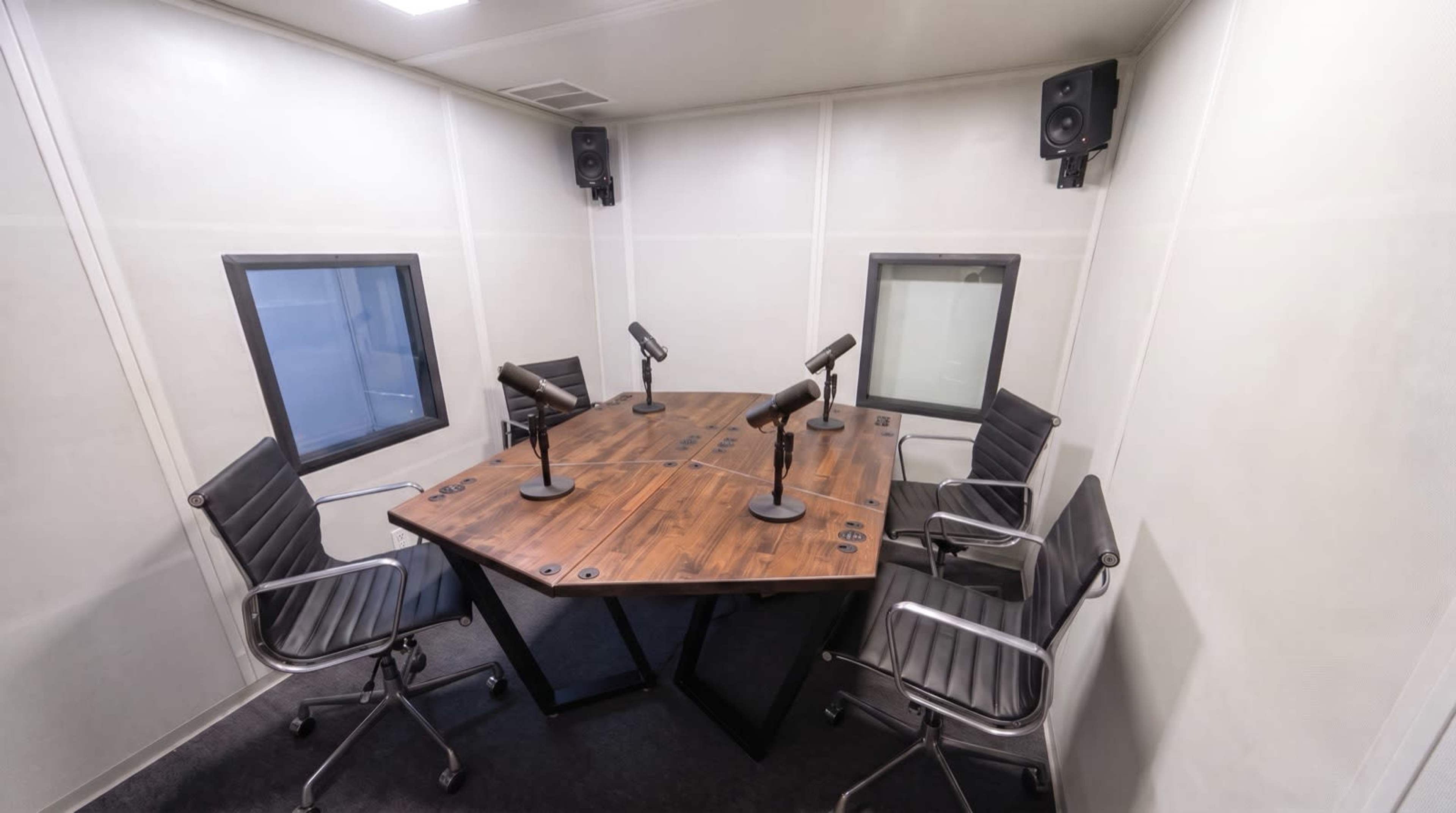 The image shows a small, soundproof recording studio with a central wooden table surrounded by four metallic chairs and microphones.