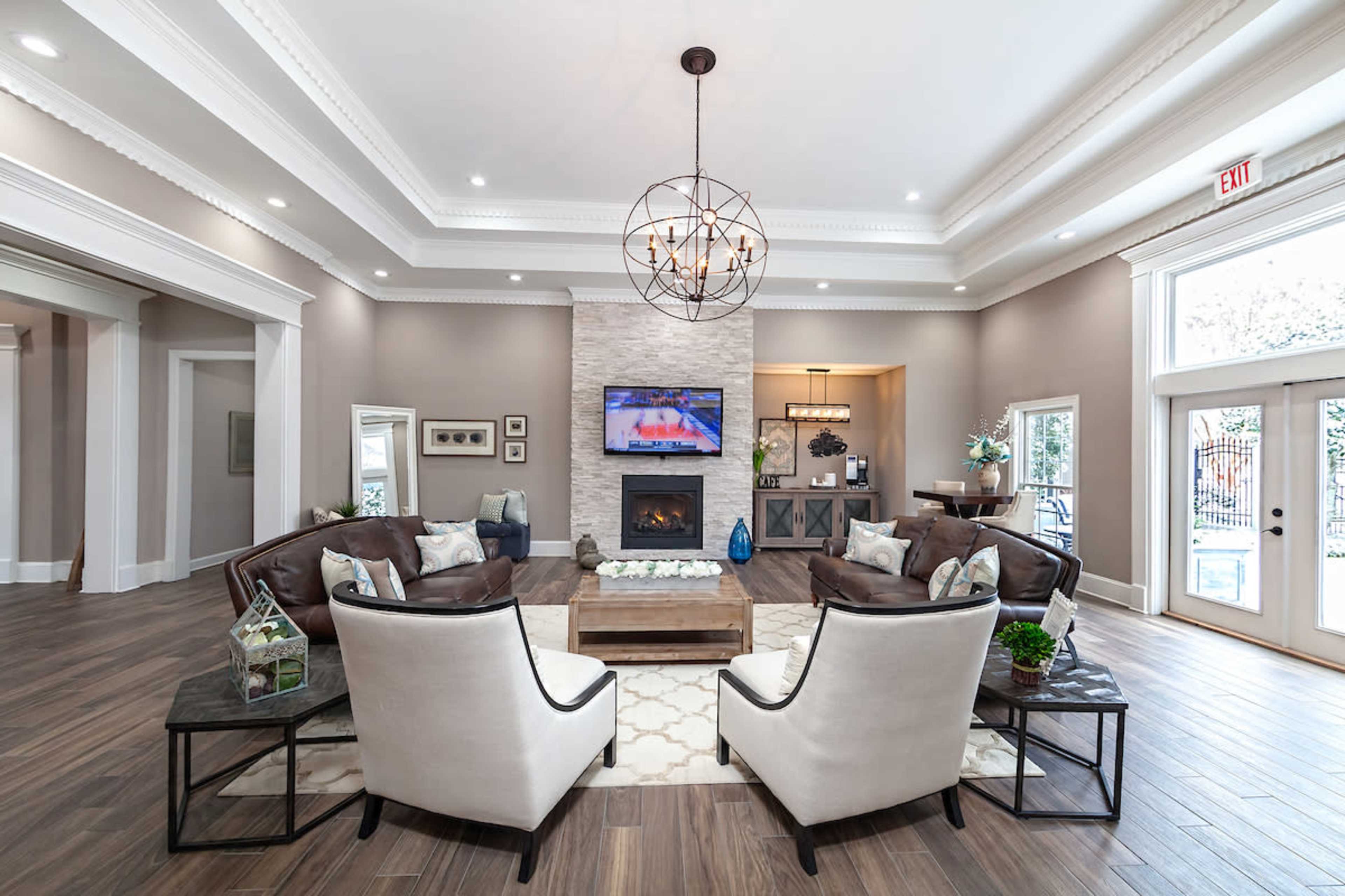 A spacious living area with a modern design featuring brown leather sofas, two cream-colored armchairs, a wooden coffee table, and a stone fireplace, all illuminated by a decorative chandelier.