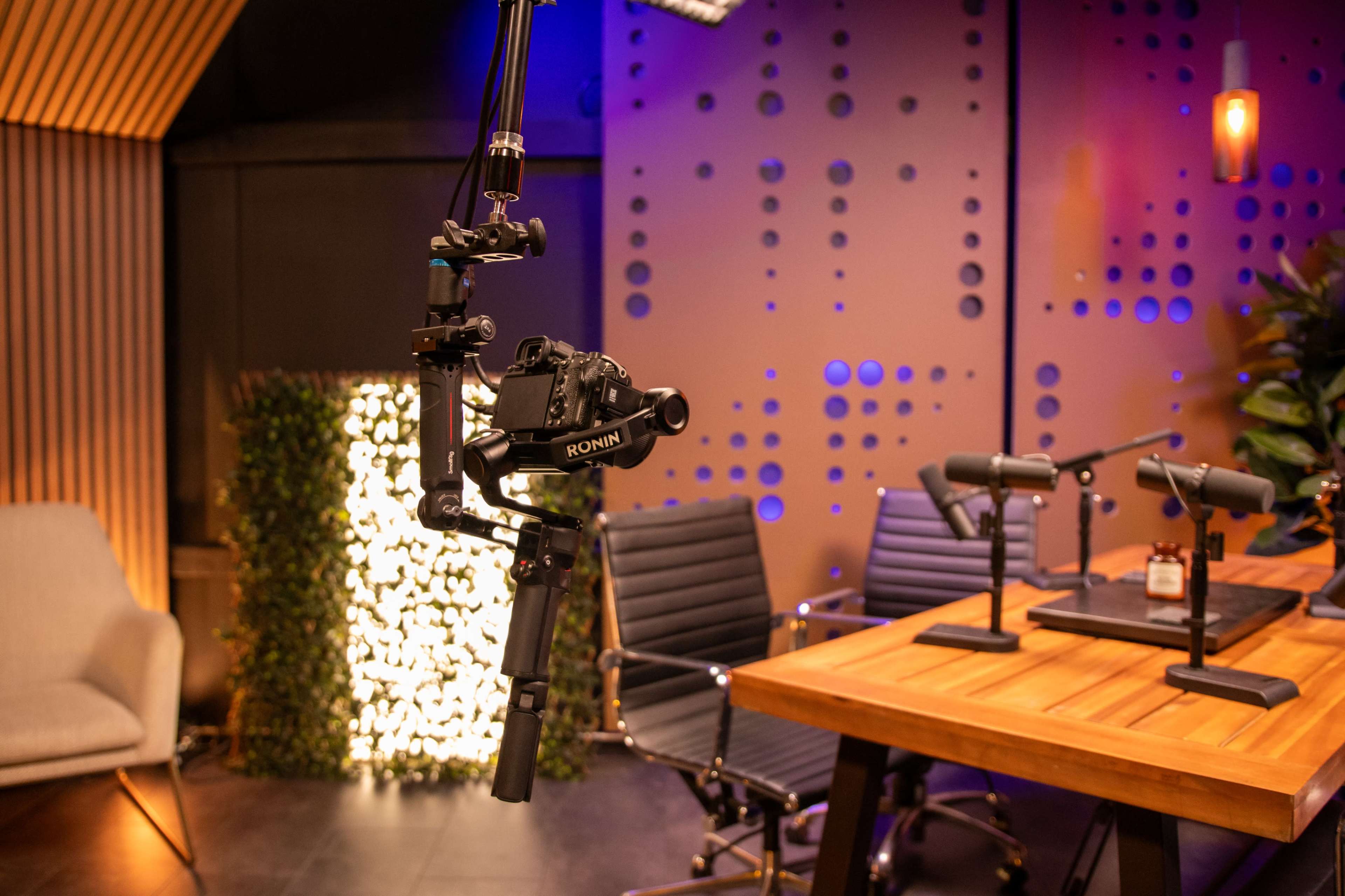 A modern studio setup featuring a camera mounted on a boom arm, multiple microphones on a table, and a backdrop with patterned walls and greenery.