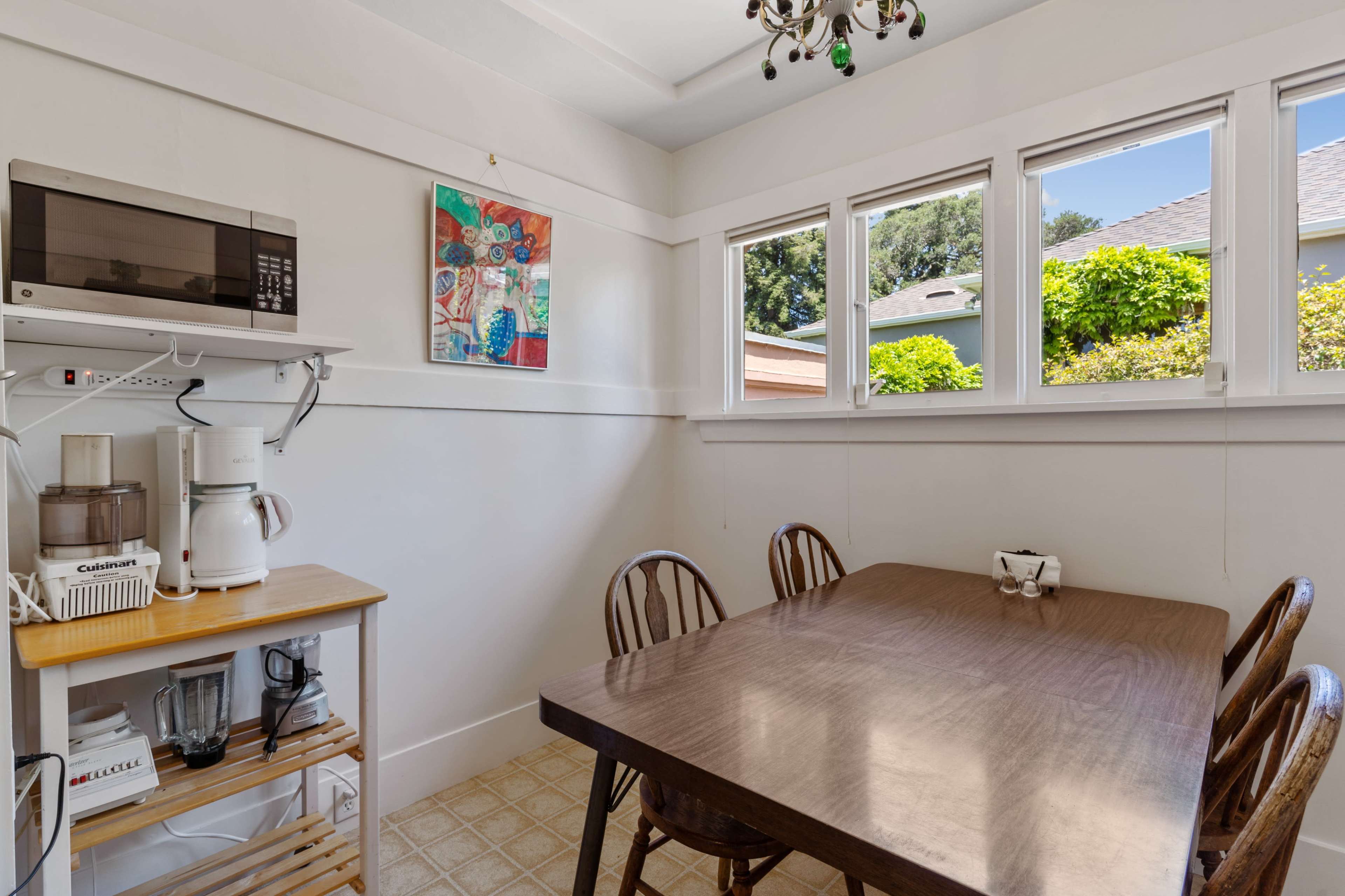 A small kitchen features a wooden dining table and chairs, with a microwave and appliances on a shelf, and windows providing natural light.